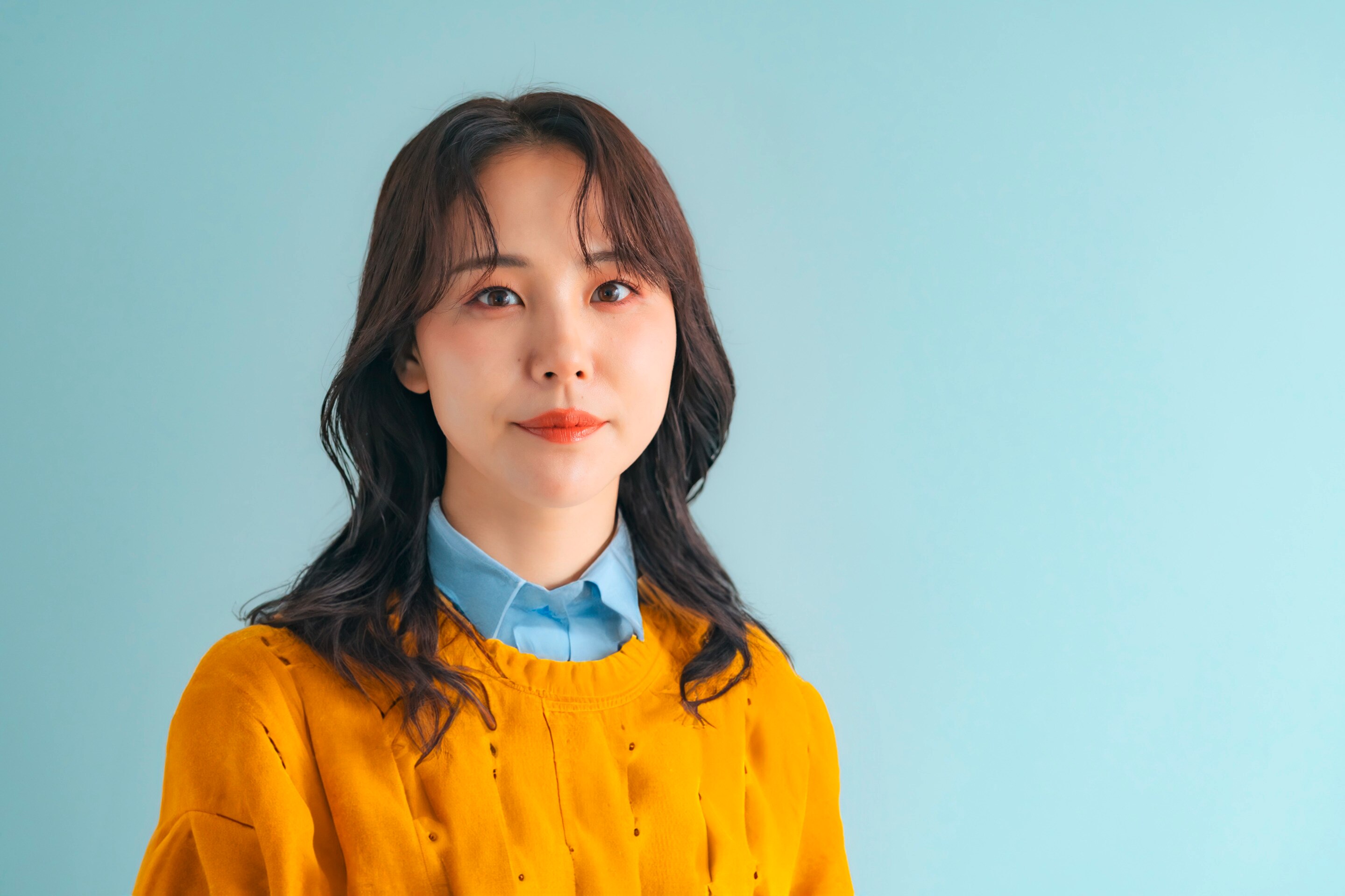 ผู้หญิงเอเชีย หน้าตรง ผมหยักศก ผมยาวเลยบ่า แสกกลาง ปอยผมม้าปัดออกข้าง ผมหน้าม้าม่านบาง สวมเชิ้ตสีฟ้า สเว็ตเตอร์สีเหลือง
