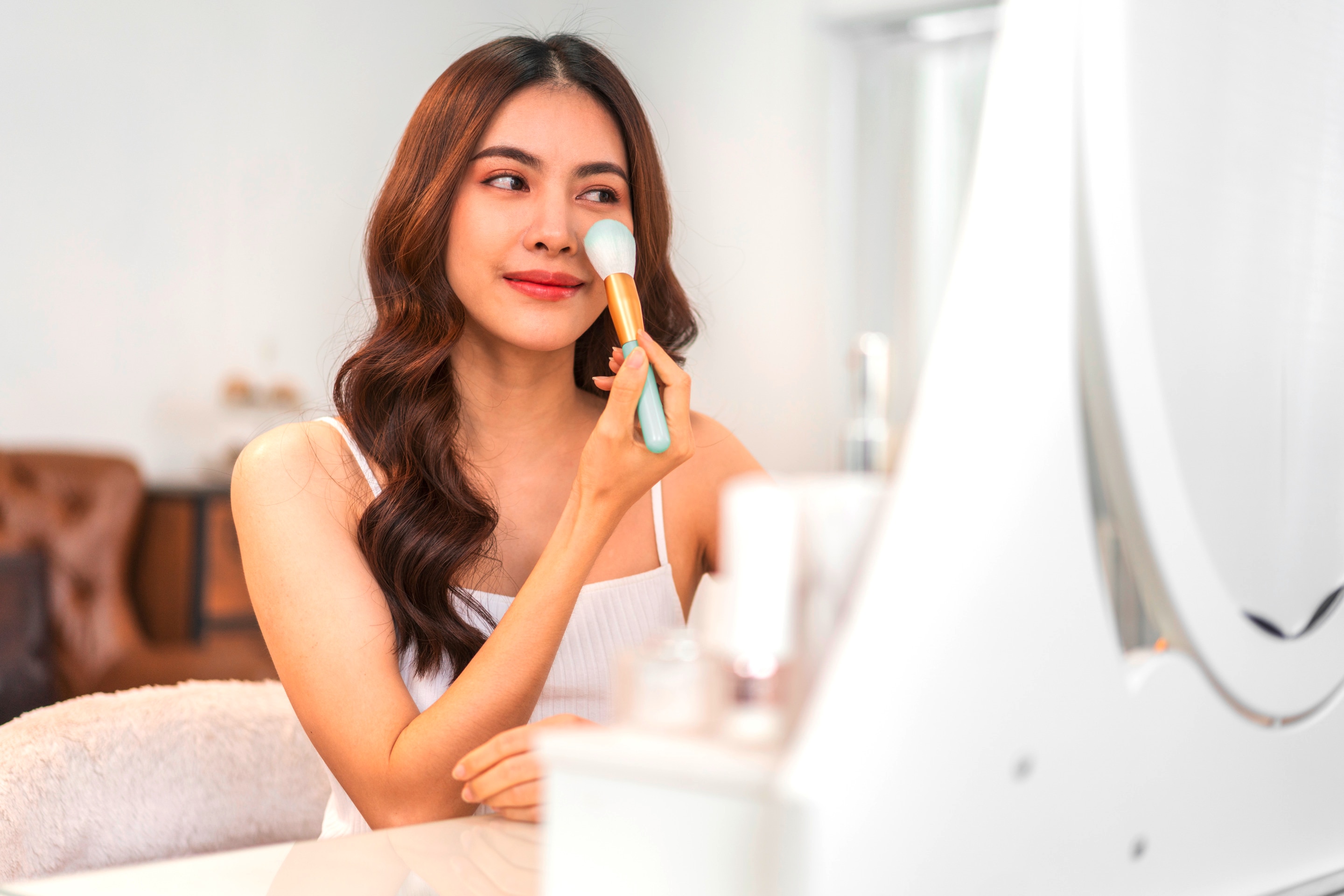 ผู้หญิงเอเชีย ผมยาว ผมหยักศก ผมดัดลอน แสกกลาง นั่งหน้าโต๊ะเครื่องแป้ง ตามองกระจก ถือแปรงปัดที่แก้ม กำลังแต่งหน้า ใส่สายเดี่ยวสีขาว