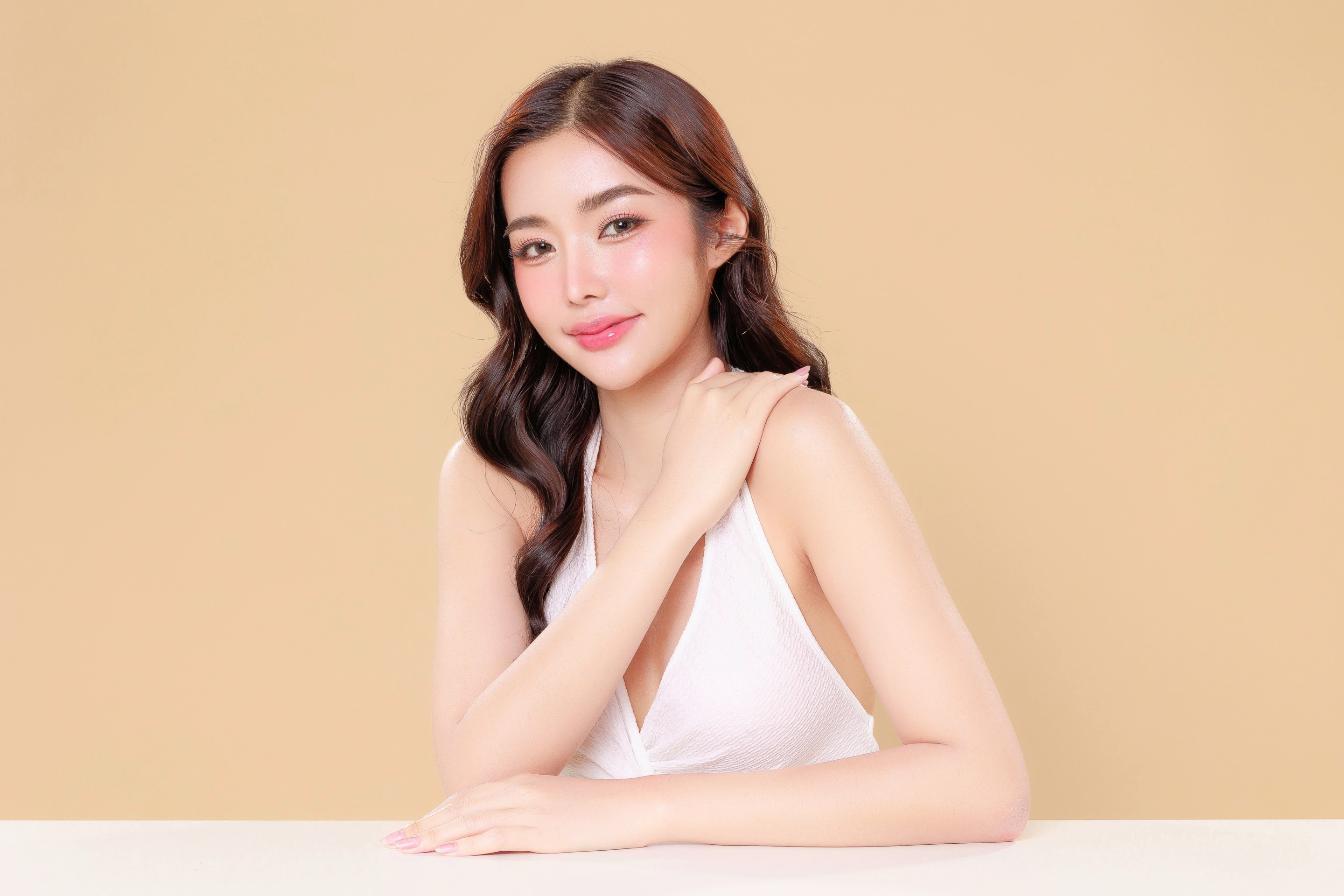 ผู้หญิงเอเชีย ผิวขาวใส ผมยาวดัดลอน แต่งหน้าโทนสีชมพู มือข้างนึงวางบนโต๊ะขาว มืออีกข้างจับบ่า สวมเดรสสีขาว