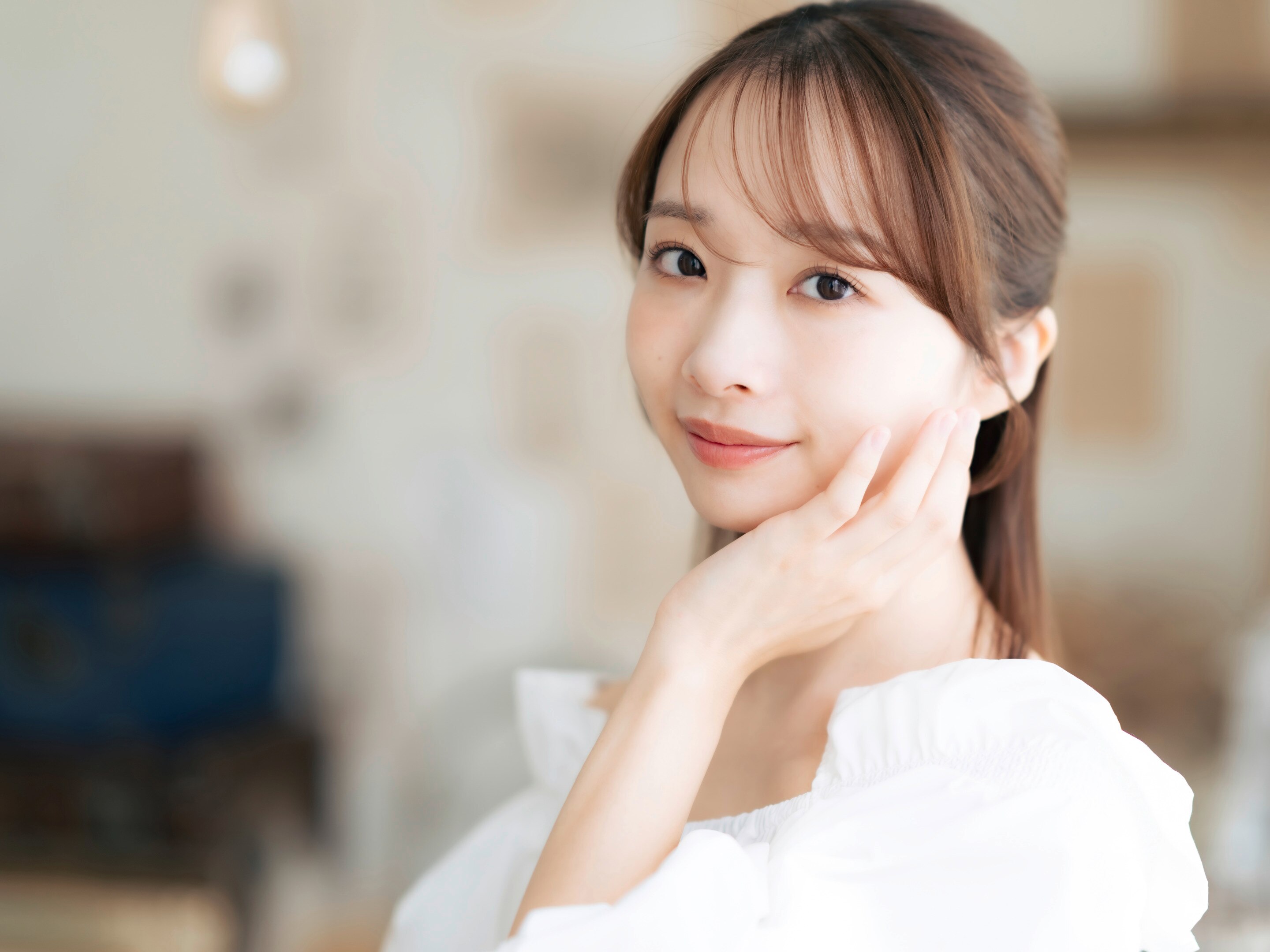 ผู้หญิงเอเชีย รวบผมครึ่งศีรษะ ผมหน้าม้าบาง ผิวสวย มือข้างหนึ่งเท้าใบหน้า สวมเสื้อสีขาว แบ็คกราวน์เบลอ
