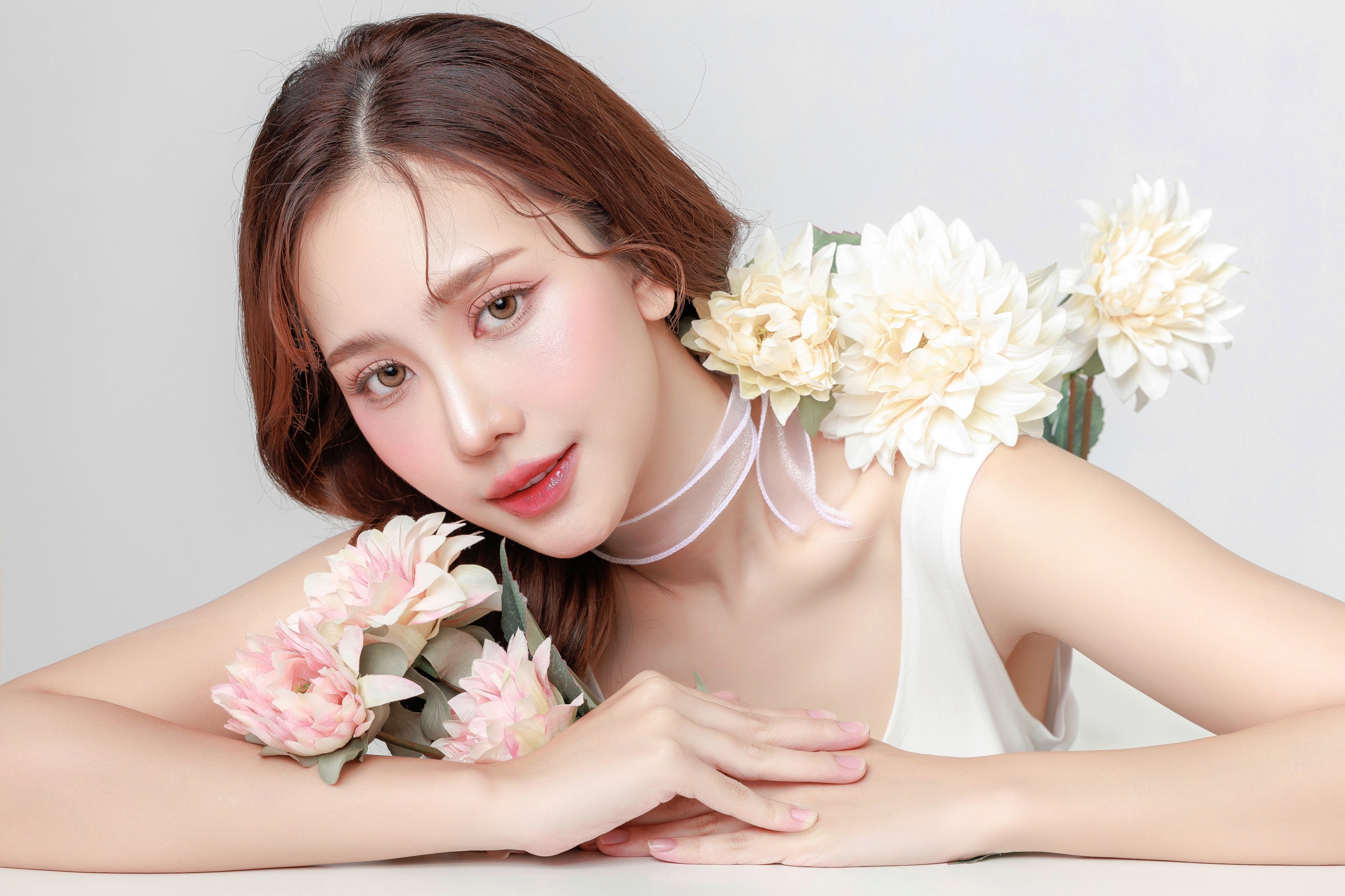 ผู้หญิงเอเชีย ผิวสวย แต่งหน้าโทนสีชมพู ผมแสกข้าง ปล่อยผม ช่อดอกไม้สีขาวที่ไหล่ ช่อดอกไม้สีชมพูใกล้หน้า มือวางประสานบนโต๊ะ หน้าเอียง ผูกริบบิ้นขาวโปร่งที่คอ สวมเสื้อกล้ามขาว
