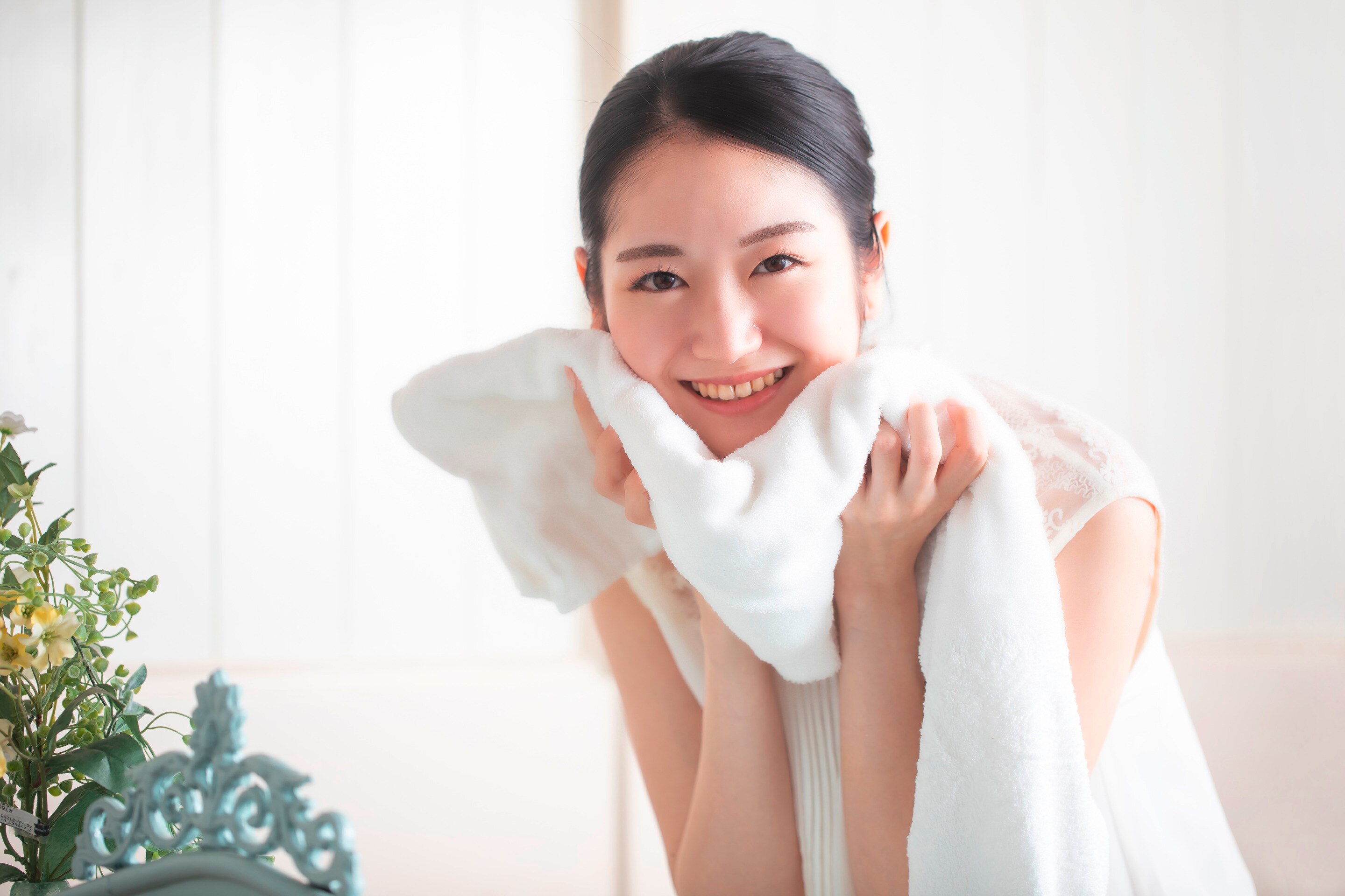 ผู้หญิงเอเชีย ผมแสกข้าง รวบผม มองตรง ยิ้มเห็นฟัน ผ้าขนหนูสีขาว สองมือยกผ้าขนหนูซับช่วงคาง สวมเสื้อแขนกุดสีขาว