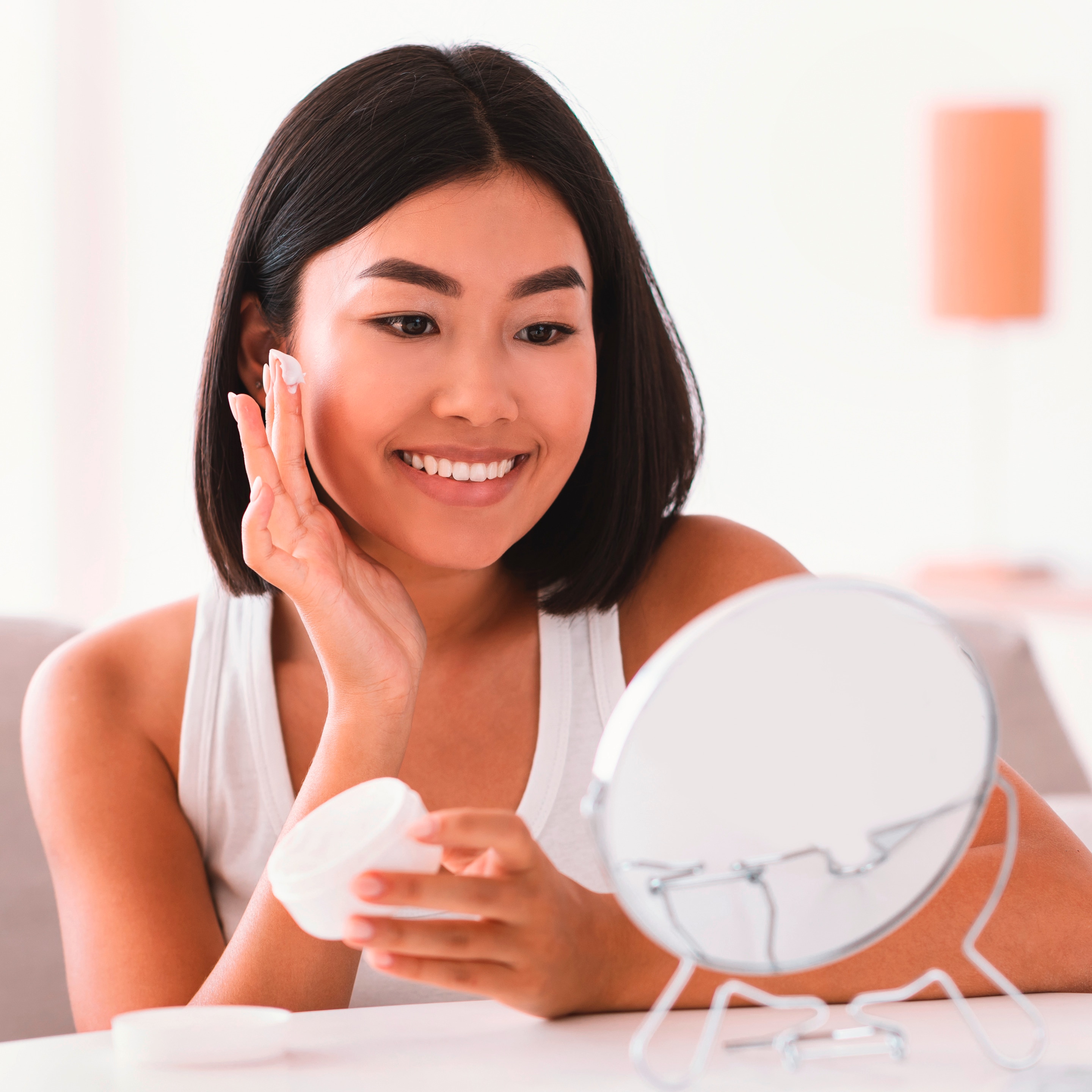 ผู้หญิงเอเชีย ผมสั้น แสกกลาง นั่งหน้าโต๊ะสีขาว กระจกตั้งโต๊ะทรงกลม ตามองกระจก ยิ้มเห็นฟัน มือหนึ่งถือกระปุกครีมเปิดฝา มือหนึ่งใช้นิ้วที่มีเนื้อครีม กำลังจะแตะที่แก้ม สวมเสื้อกล้ามสีขาว มีขวดสกินแคร์ใสวางบนโต๊ะ