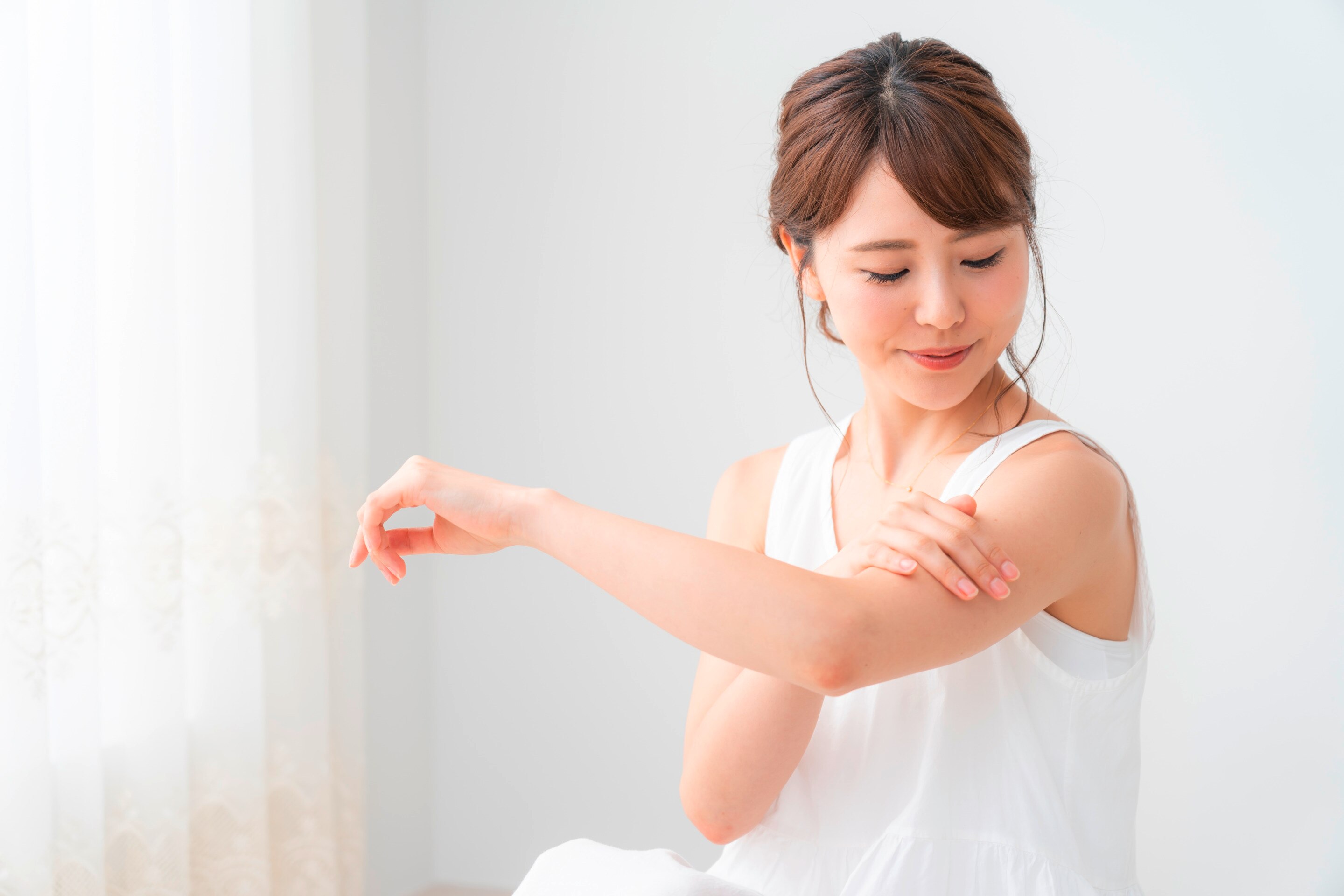 ผู้หญิงเอเชีย รวบผม ผิวสวย ตาเหลือบมองต้นแขนที่ใช้มือสัมผัส สวมชุดแขนกุดสีขาว