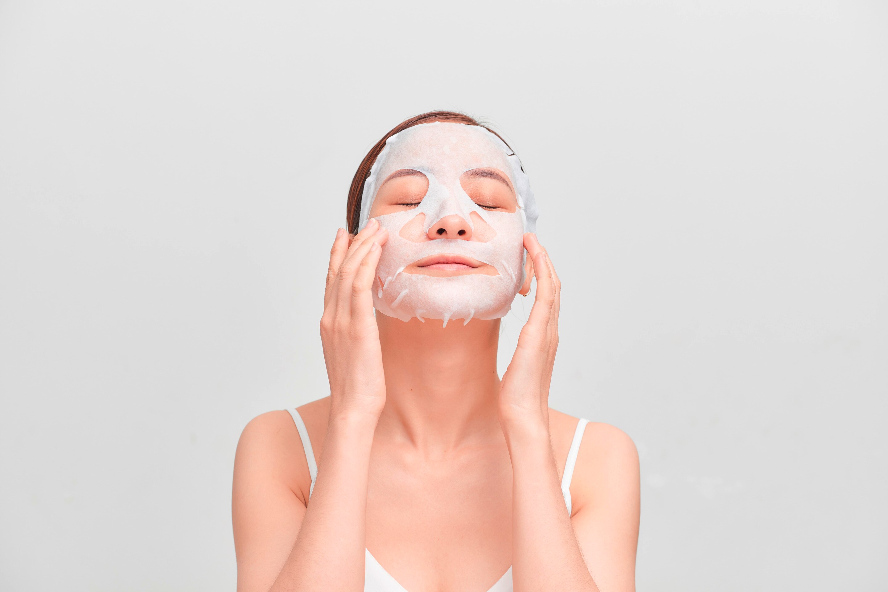 ผู้หญิงแปะแผ่นมาส์กบนหน้า หลับตา เงยหน้าเล็กน้อย ใช้สองมือแตะแผนมาส์กบนหน้า ใส่เสื้อสายเดี่ยวสีขาว