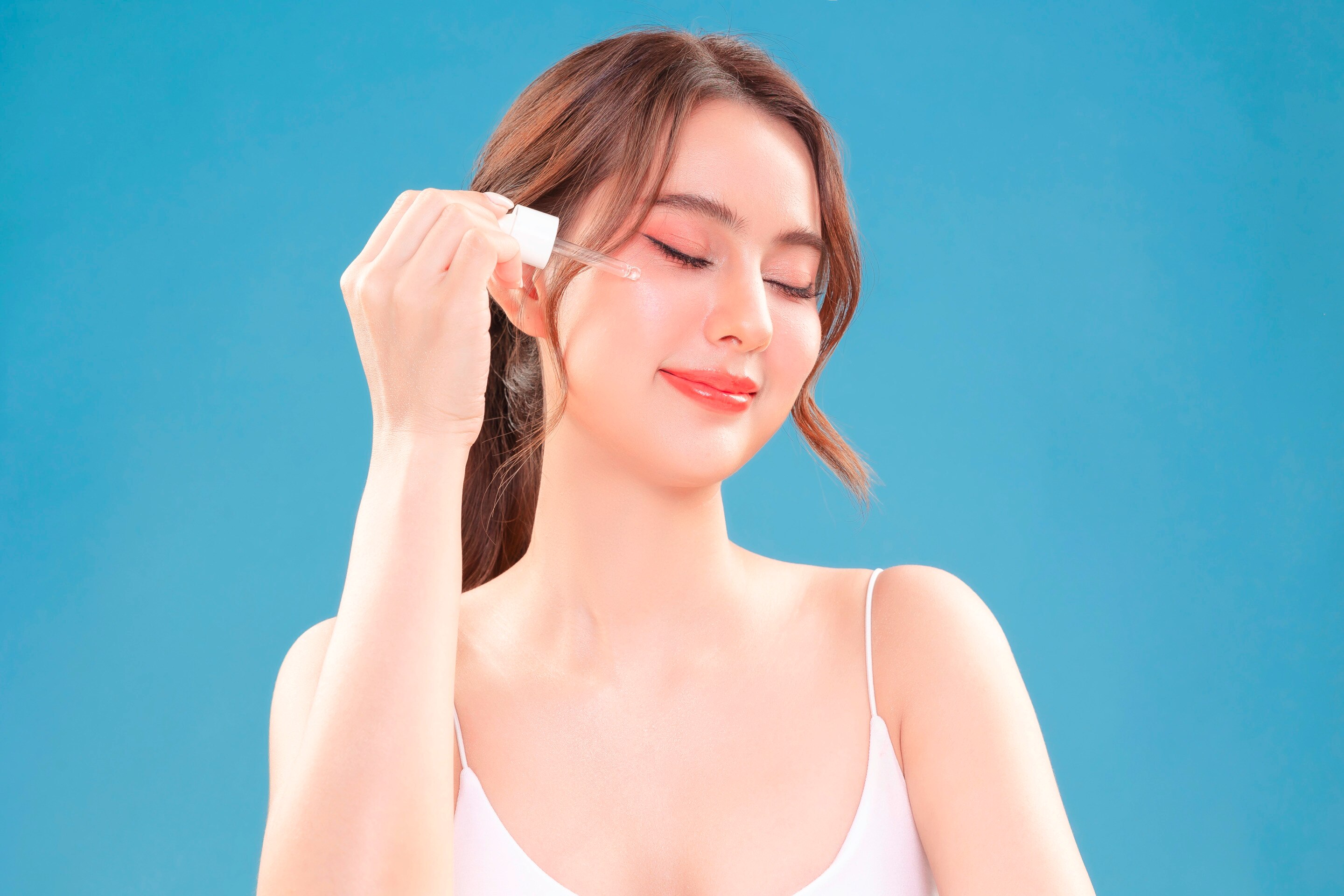 ผู้หญิงเอเชีย ผมยาวรวบไว้ด้านหลัง หลับตา ถือดรอปเปอร์หยดเซรั่มบนหน้า สวมเสื้อสายเดี่ยวสีขาว