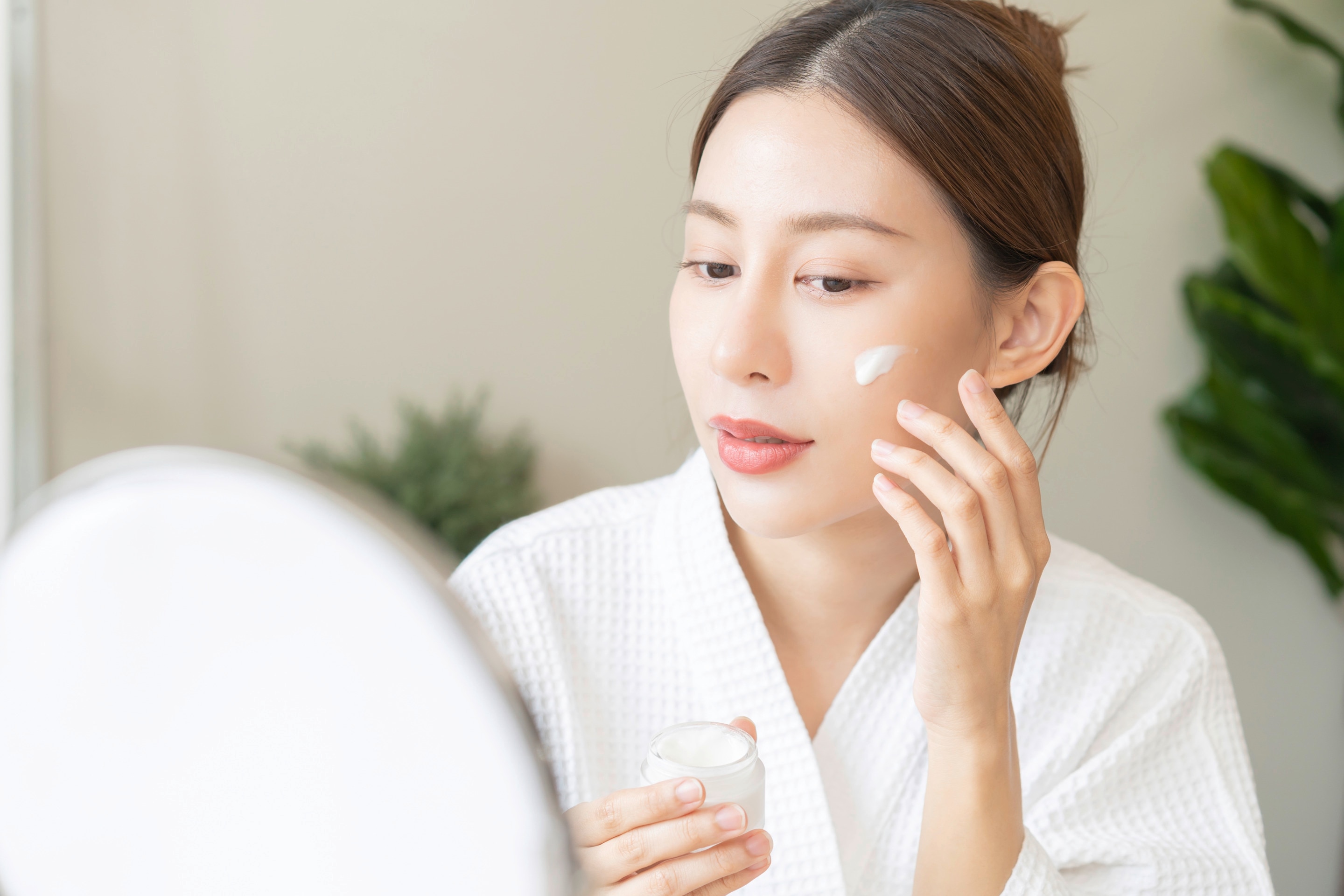 ผู้หญิงเอเชีย รวบผม สวมเสื้อคลุมอาบน้ำ มือนึงถือกระปุกครีมสีขาว อีกมือแตะใบหน้าที่มีเนื้อครีมป้ายไว้ 