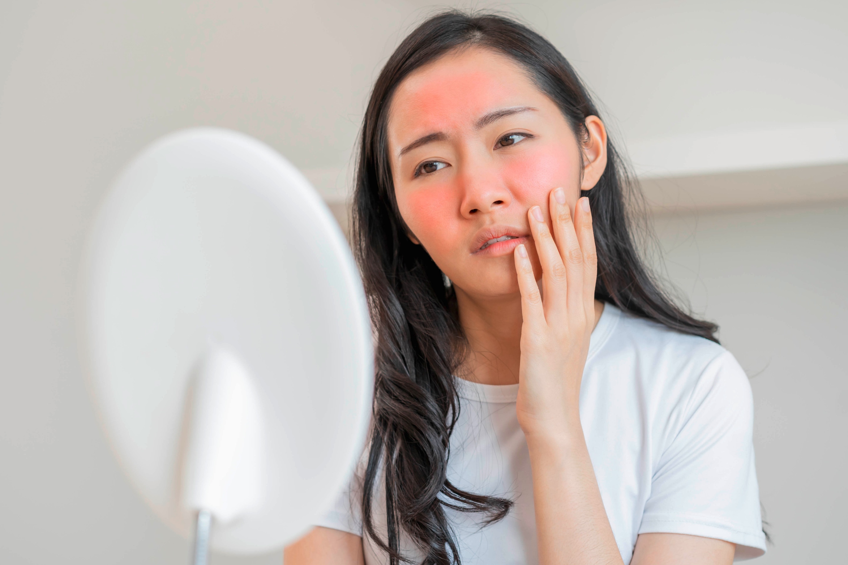 Asian woman looks at red face in a vanity mirror.