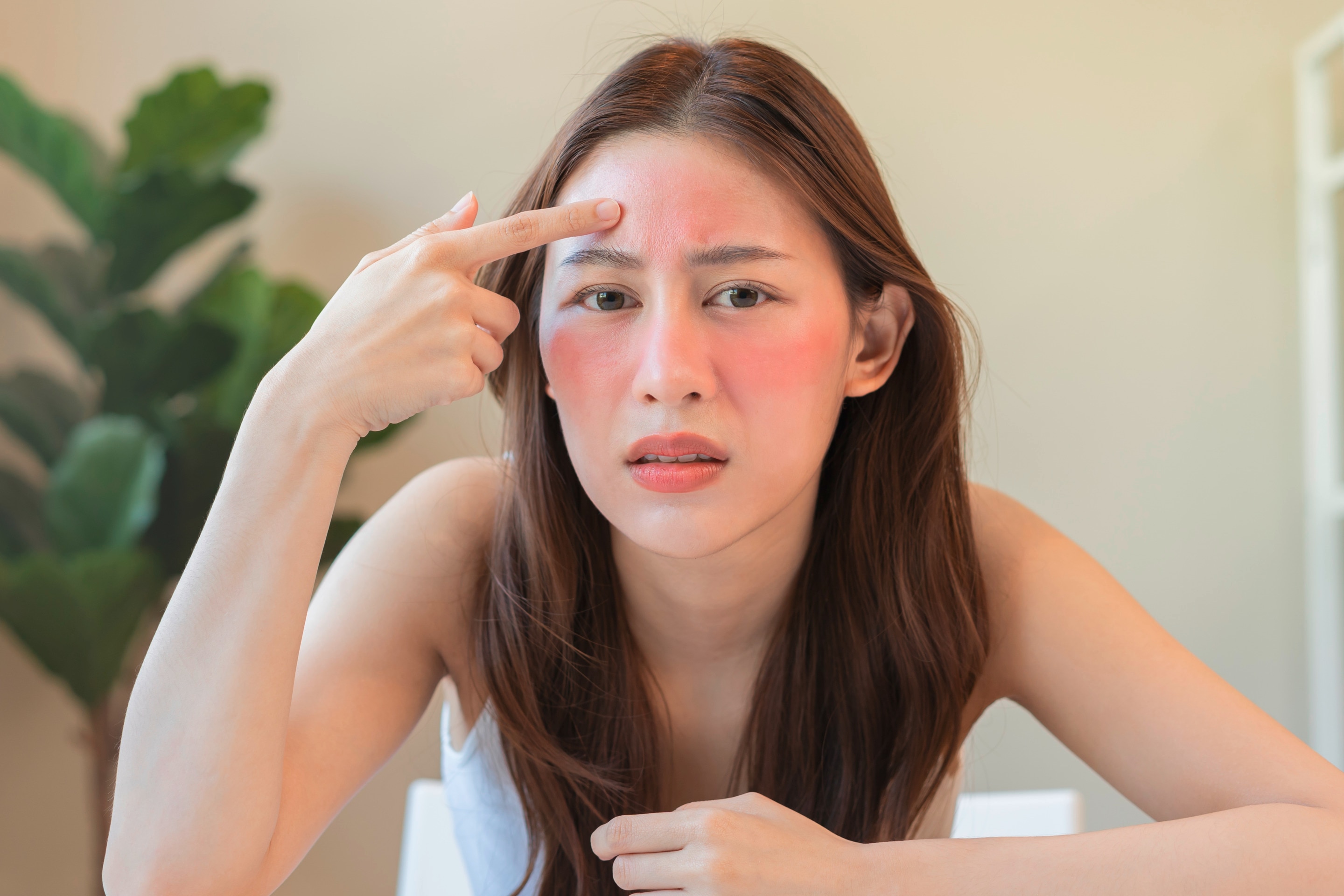 ผู้หญิงเอเชีย ผมตรงสั้น นั่งส่องกระจก ขวดผลิตภัณฑ์สีขาวหลายรูปทรงบนโต๊ะ มือถือสำลีเช็ดบนแก้ม สวมเสื้อยืดสีขาว