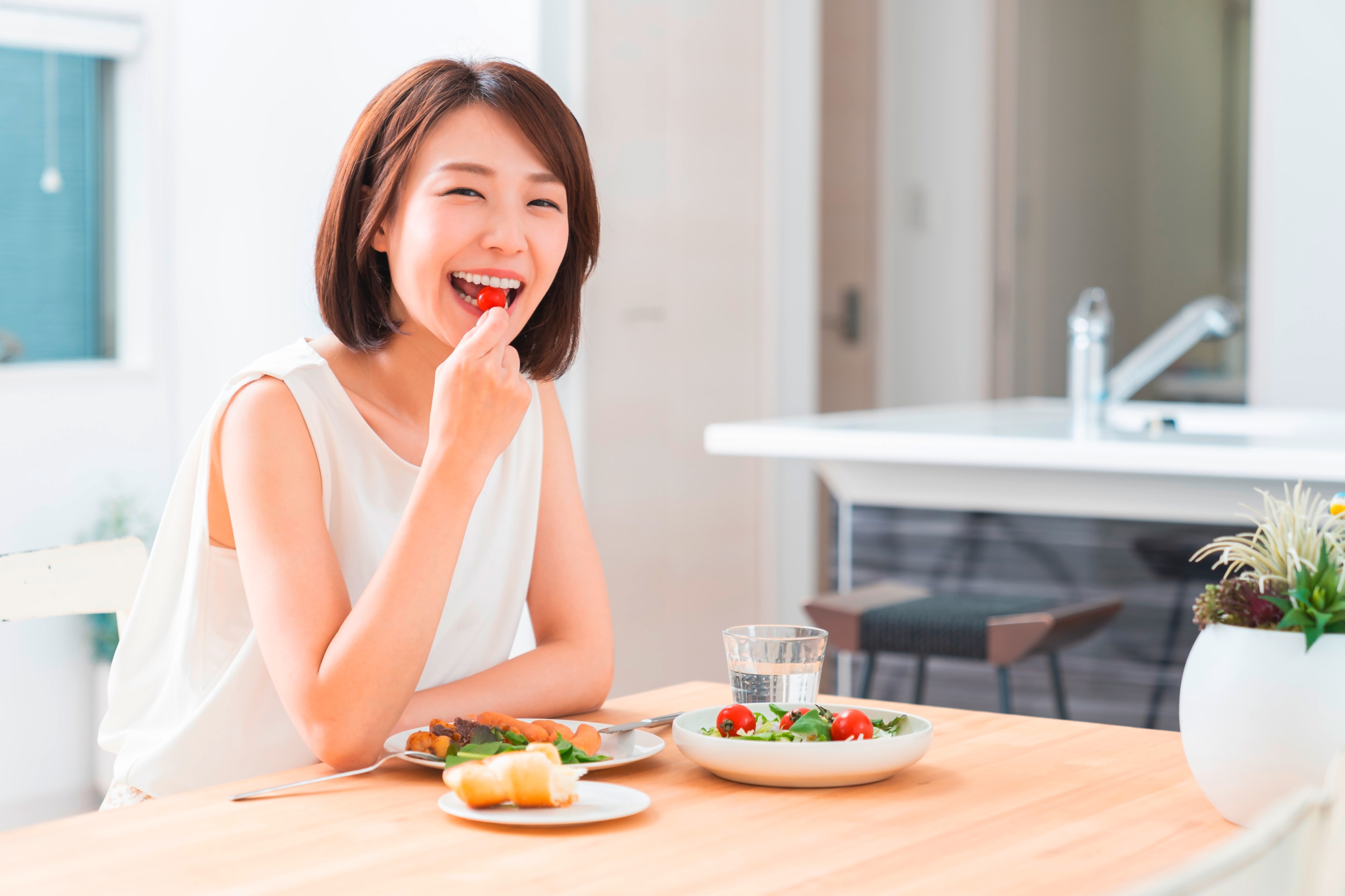 ผู้หญิงเอเชีย ผมสั้น แสกข้าง ใบหน้ายิ้ม มือหยิบมะเขือเทศลูกเล็กใส่ปาก สวมเสื้อแขนกุดสีขาว บนโต๊ะด้านหน้าวางอาหาร 3 จาน จานผักสลัด แก้วใส่น้ำเปล่า