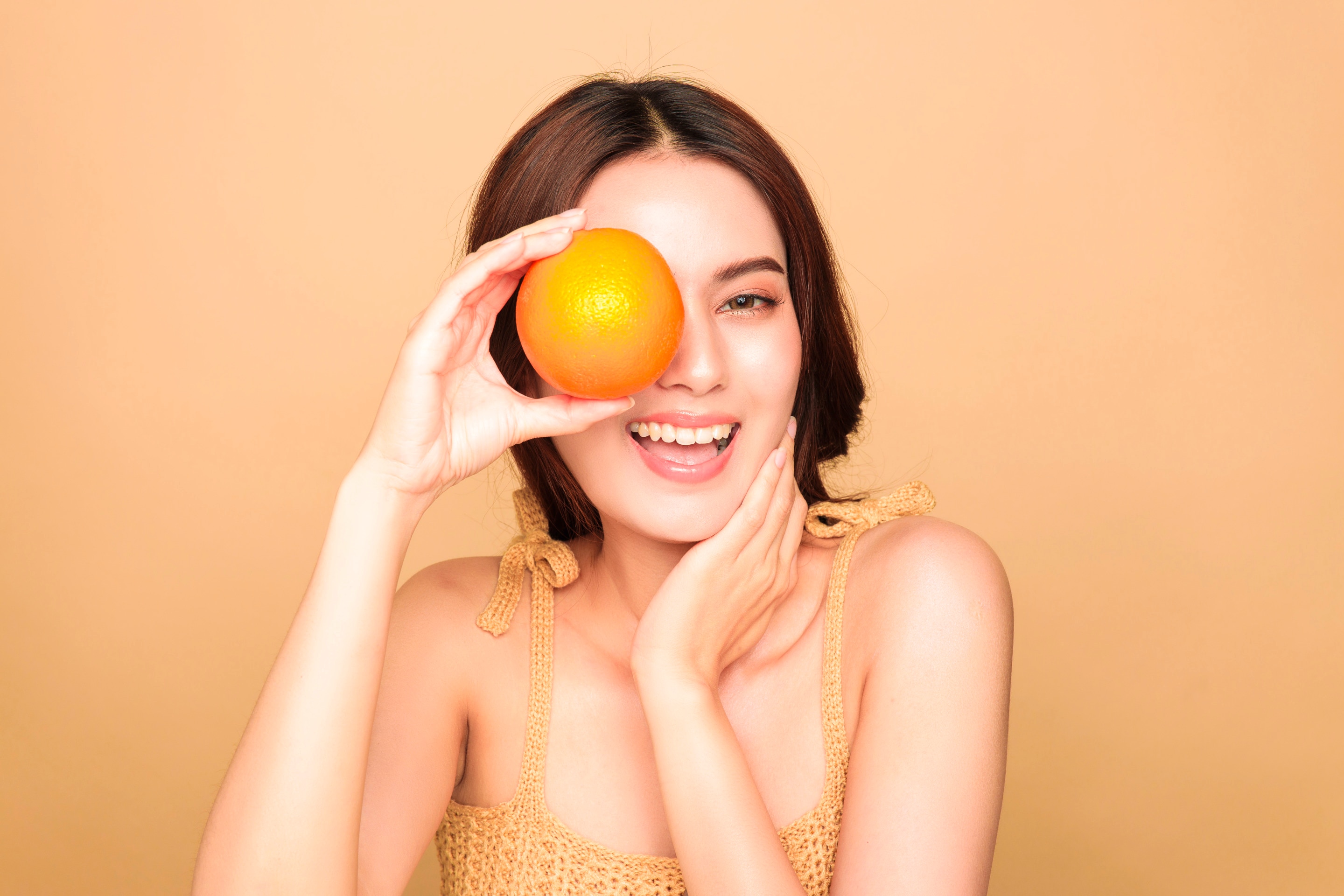 ผู้หญิงเอเชีย ยิ้มเห็นฟัน มือข้างนึงถือส้มบังช่วงดวงตา อีกมือค้ำหน้าช่วงคาง ใส่สายเดี่ยวสีเหลือง