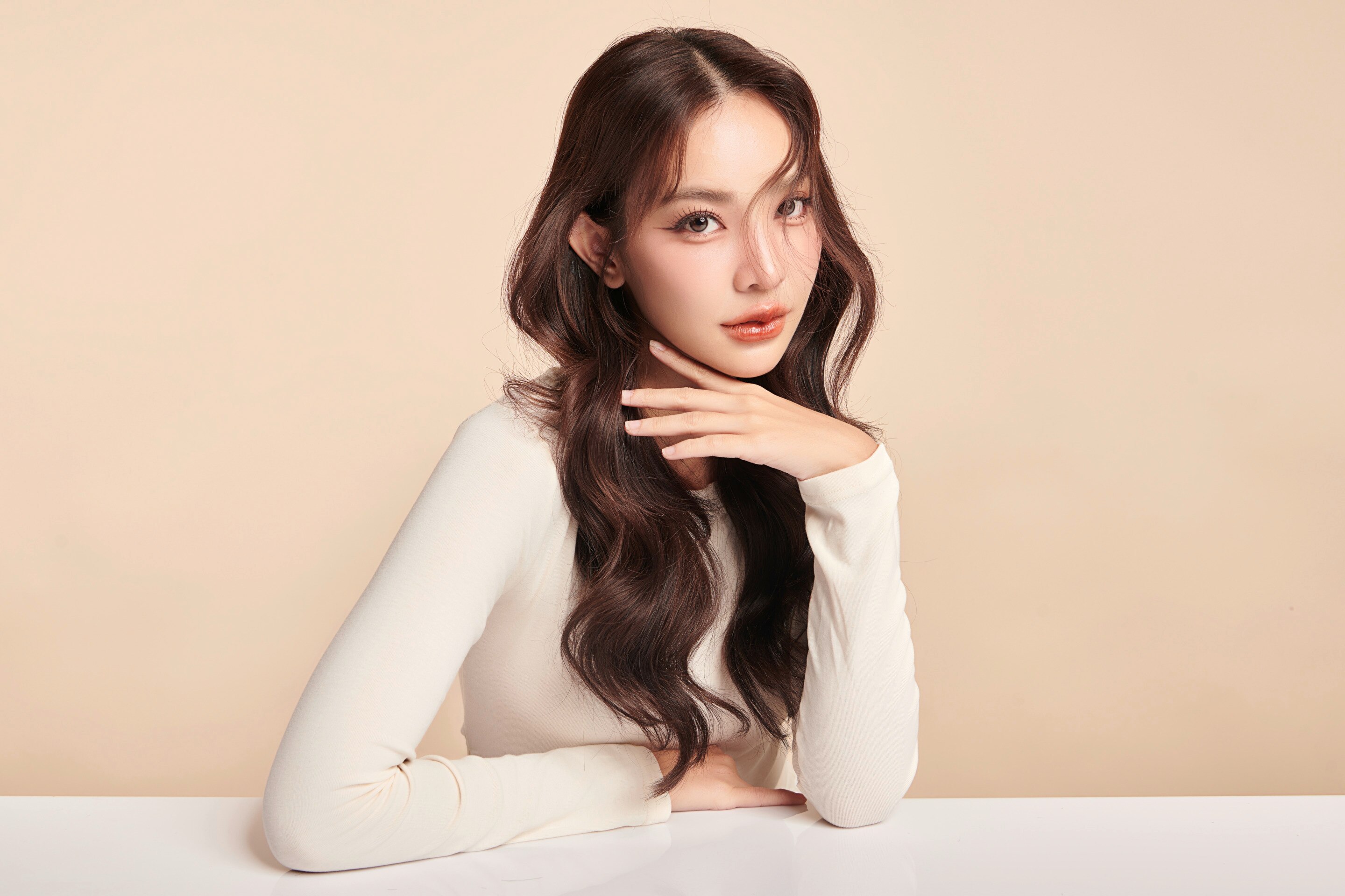 ผู้หญิงเอเชีย ผมลอนยาว ผมสไลซ์ไม่เท่ากัน หน้าเอียง เท้าแขนบนโต๊ะขาว มือนึงคว่ำแตะคาง สวมเสื้อแขนยาวสีขาว