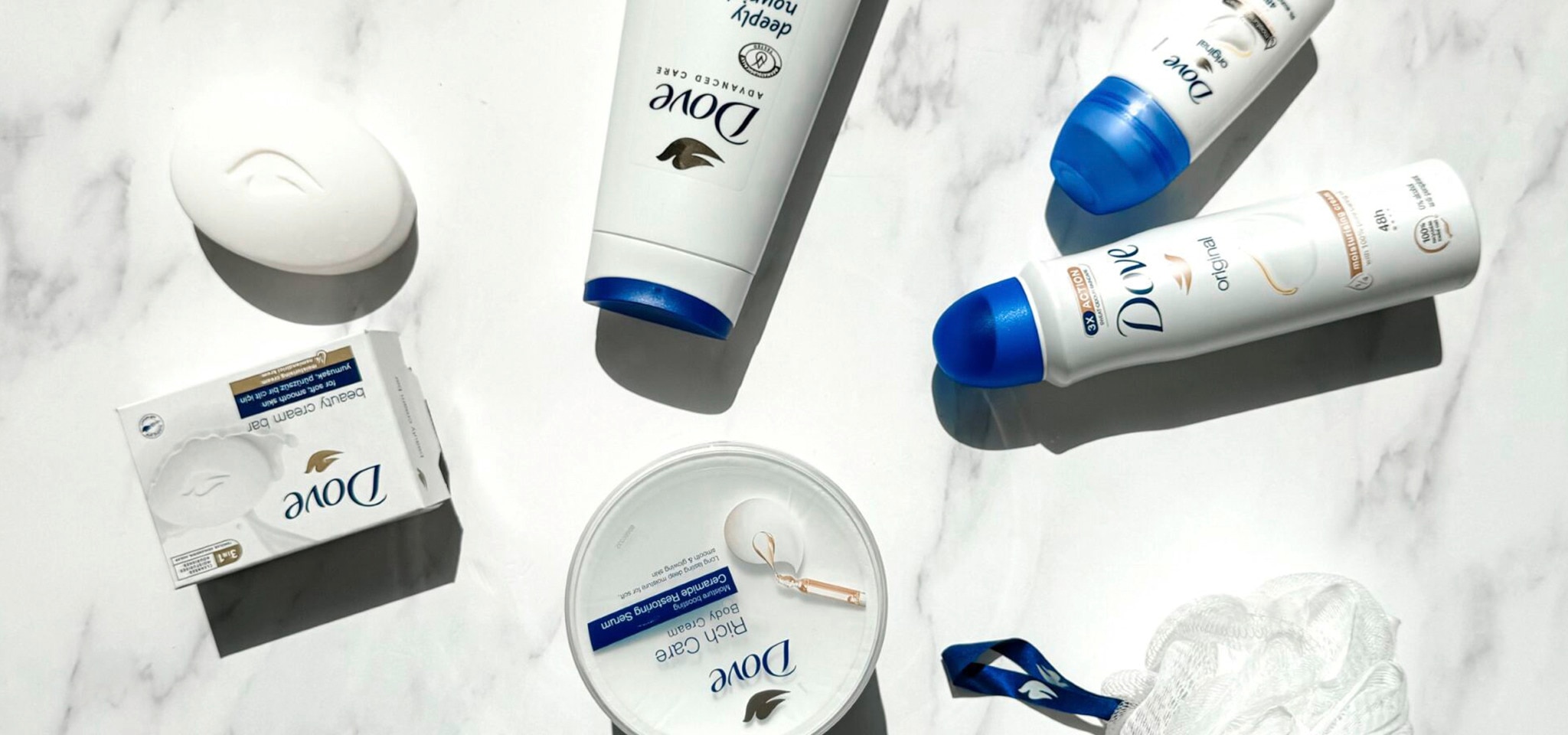 Dove Moisturising cream, beauty bar, body wash and deodorant with a loofah on a white marble surface.