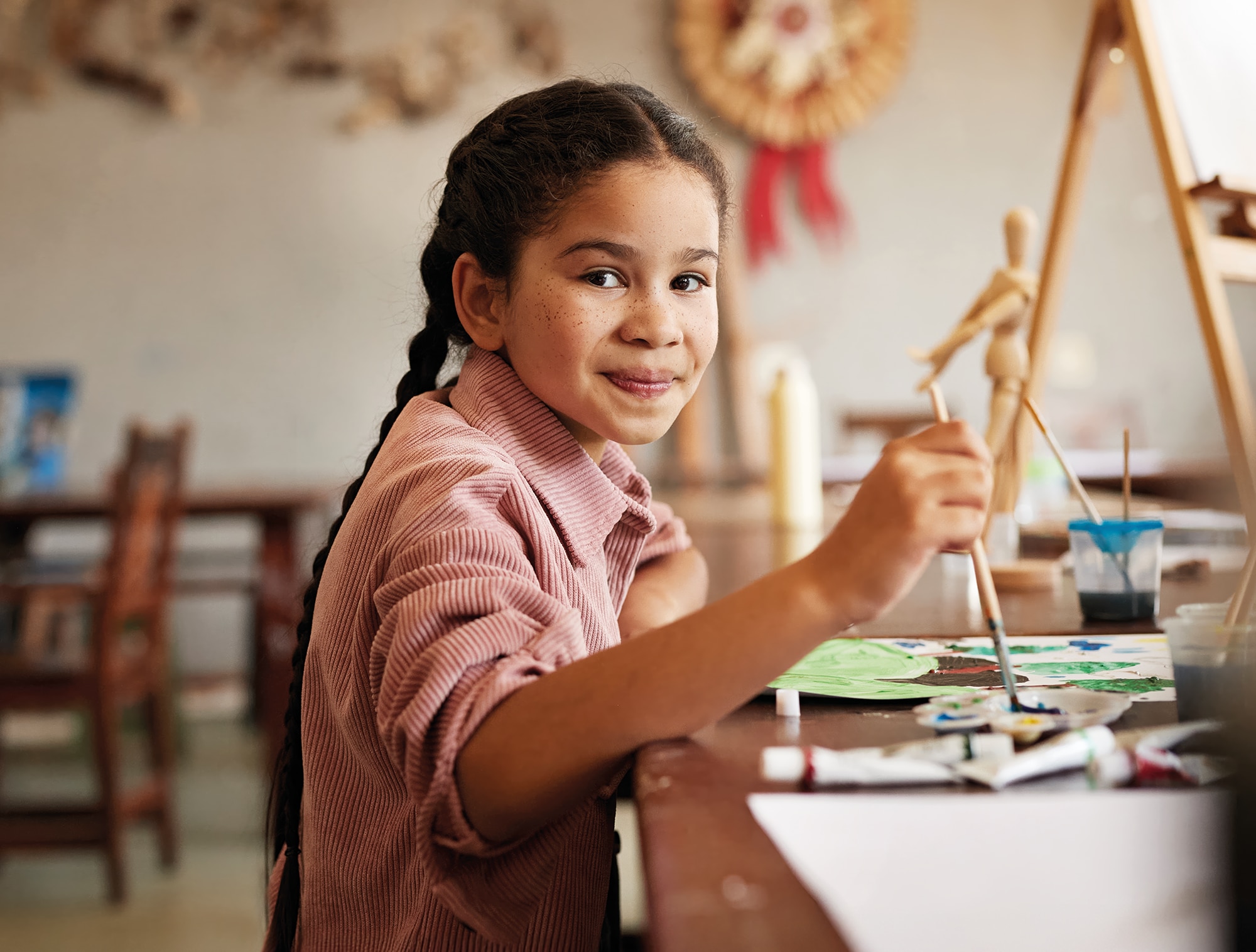 A girl sitting at a desk smiling with a paint brush in hand 