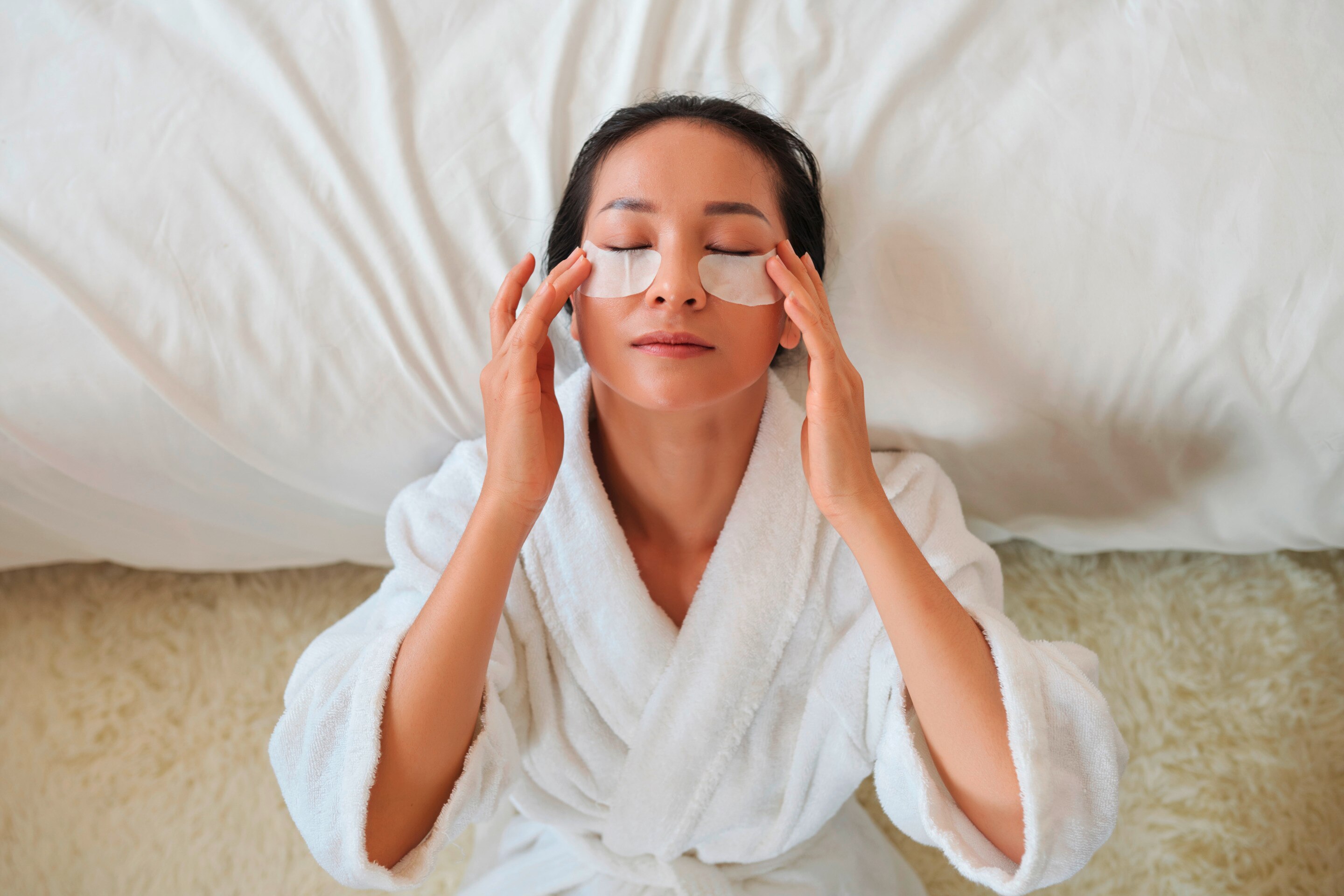 Woman in bathrobe putting on eye patches on her under-eye area.