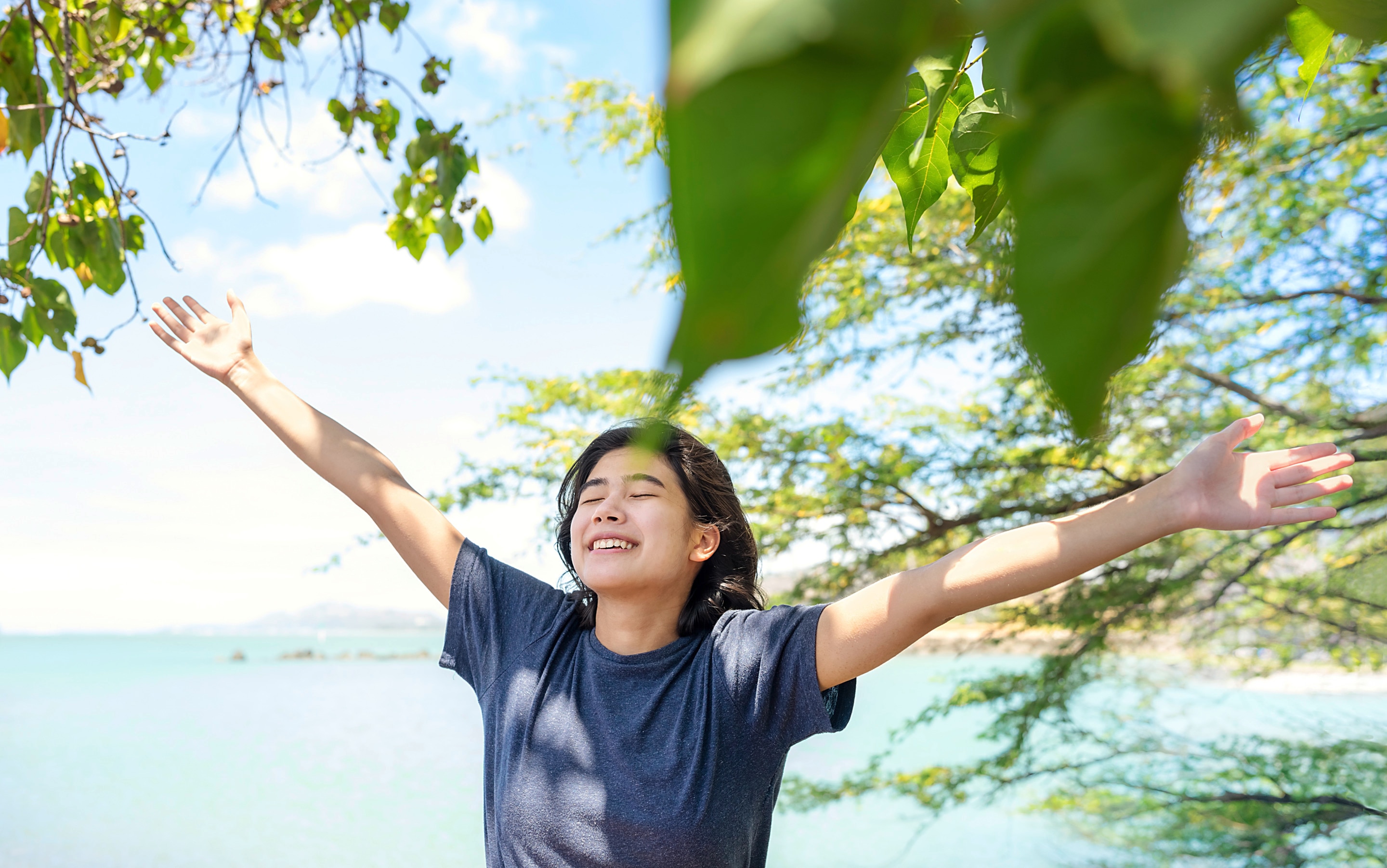 Asian girl in blue shirt with arms raised outdoors.