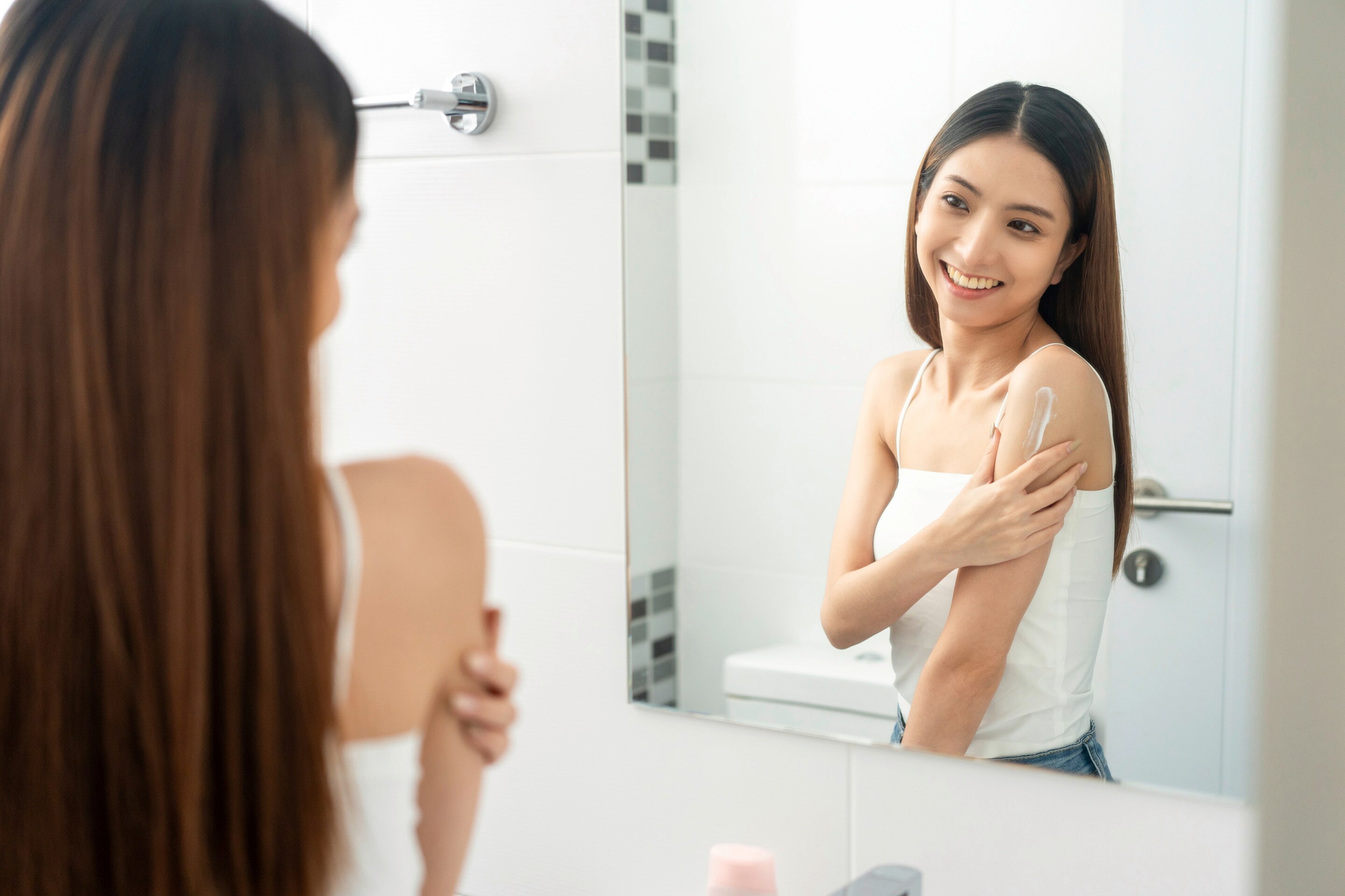 Woman rubbing lotion on her arm while standing in front of a mirror.