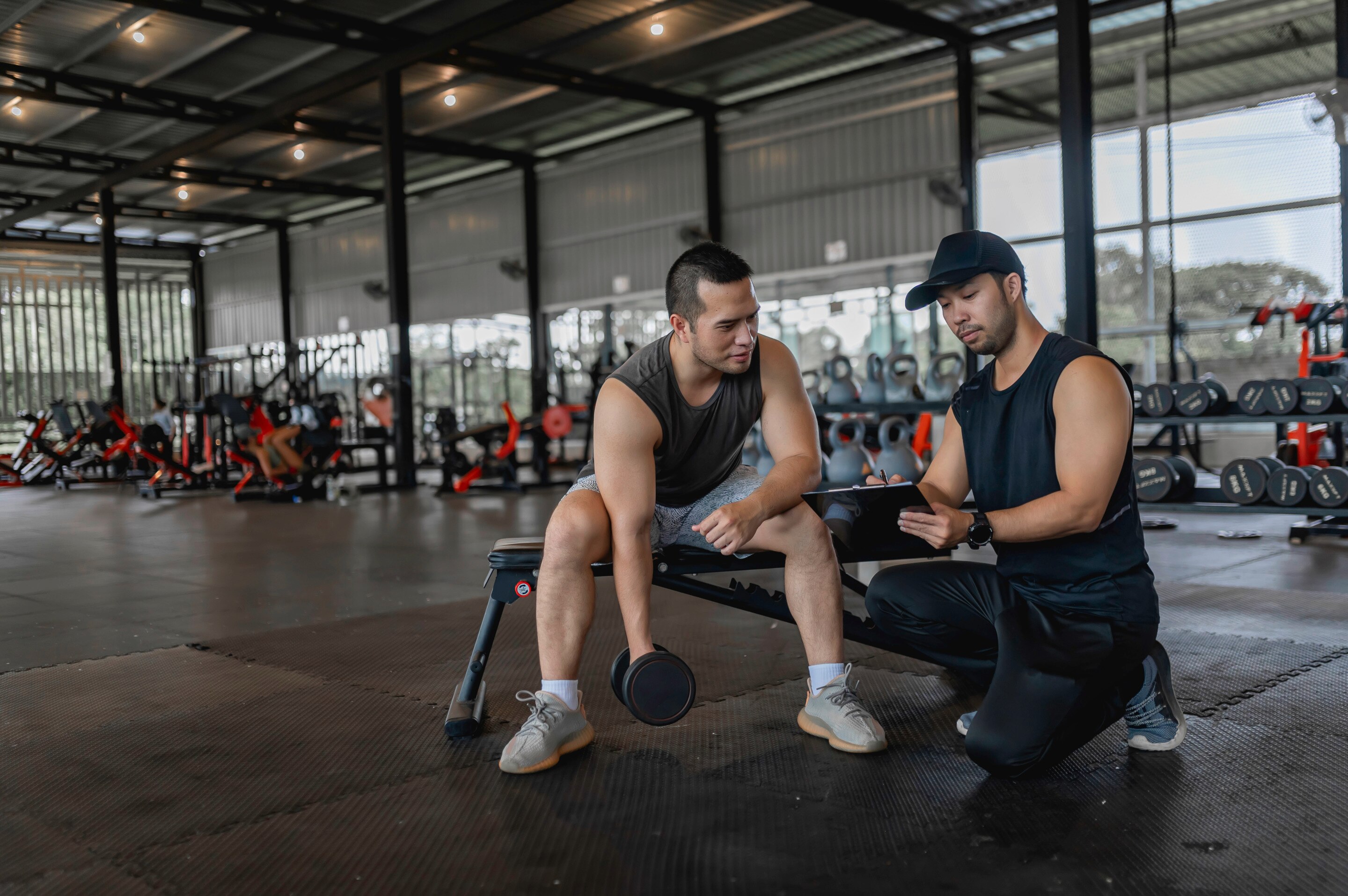 Man lifting barbell with a personal gym trainer showing him notes.