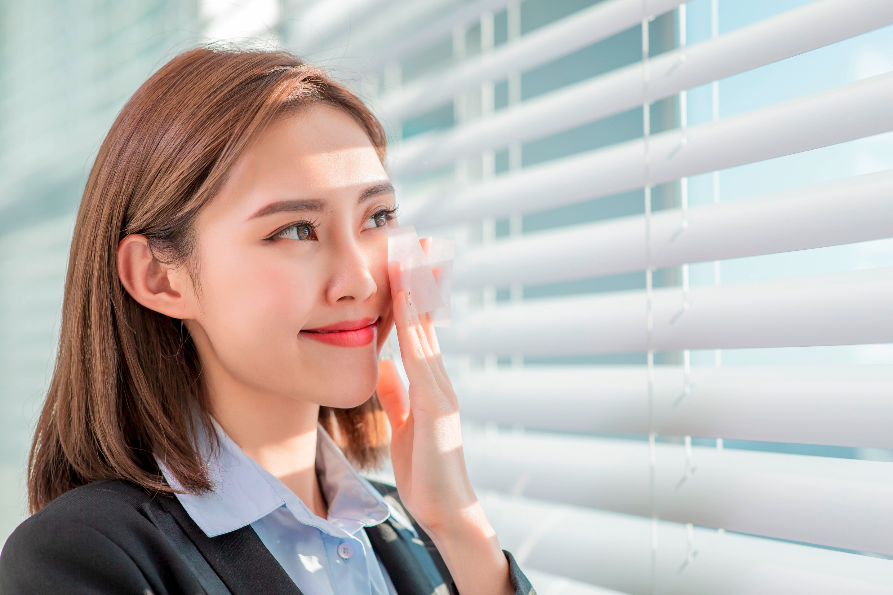 Woman in office wear using blotting paper on her cheek.