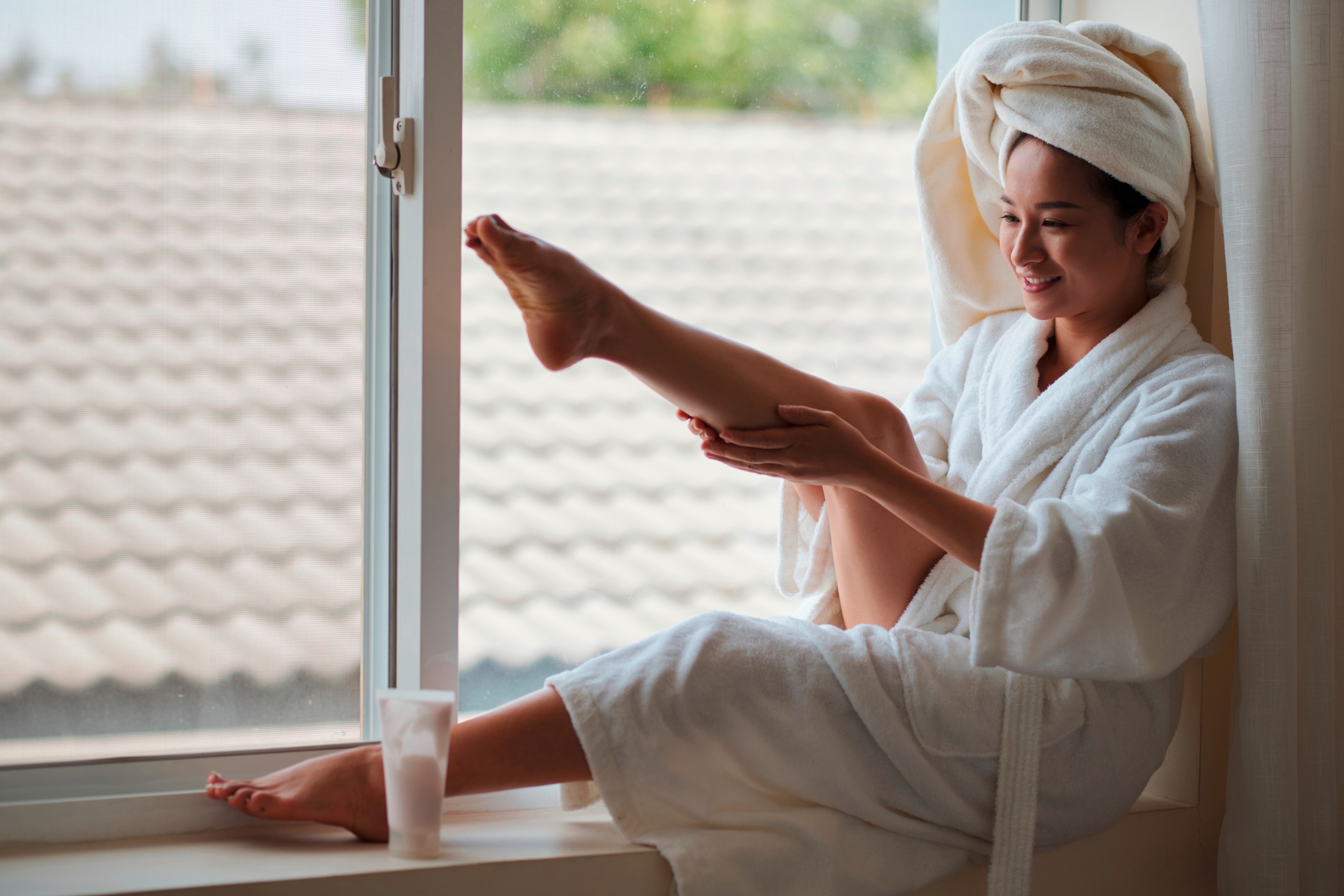 Woman in bathrobe applying lotion to her leg while sitting near a window.