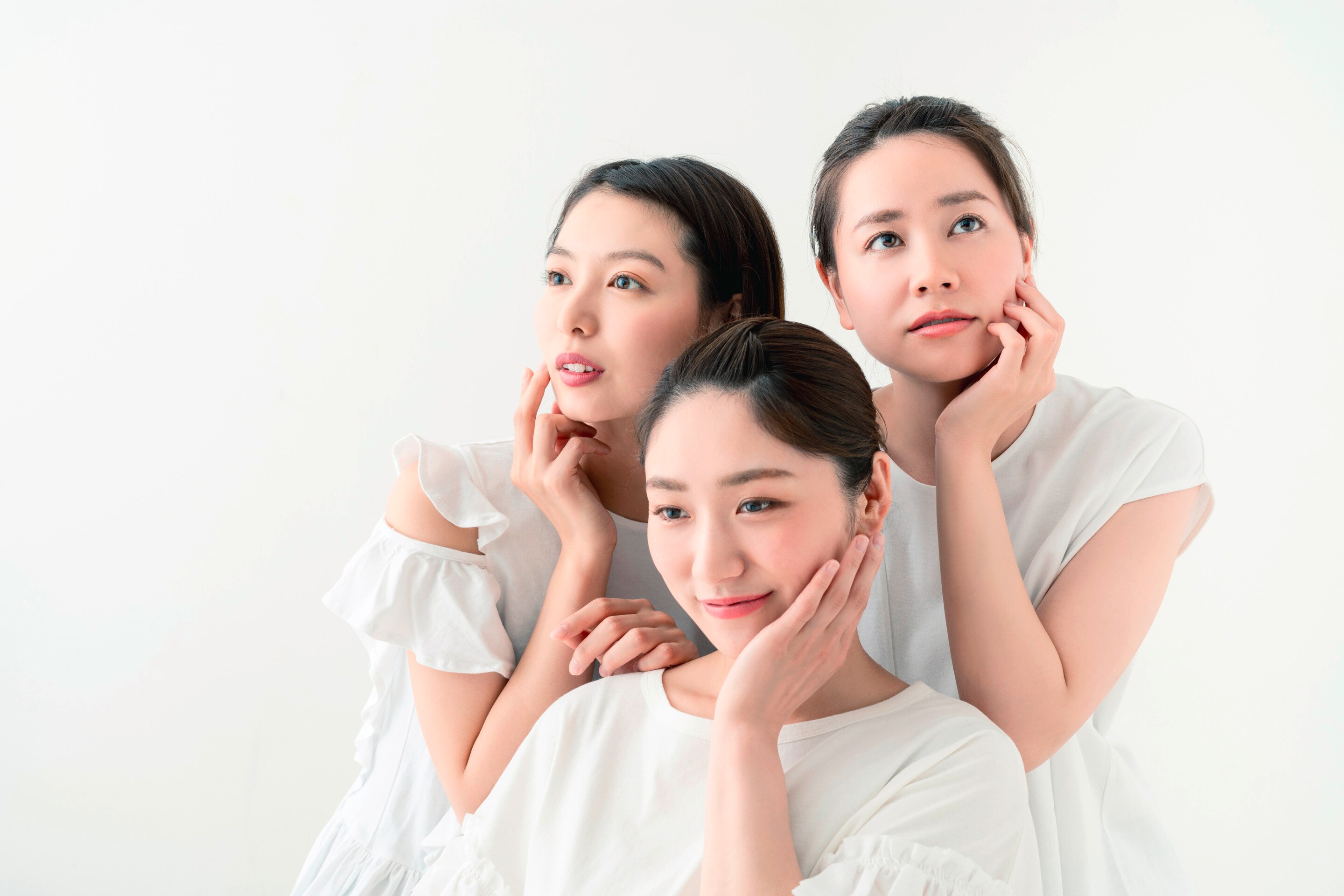 ผู้หญิงเอเชีย สามคนสามวัย คนตรงกลางนั่ง สองคนยืนด้านหลัง รวบผม ใช้มือข้างหนึ่งแตะผิวหน้า สวมชุดสีขาว