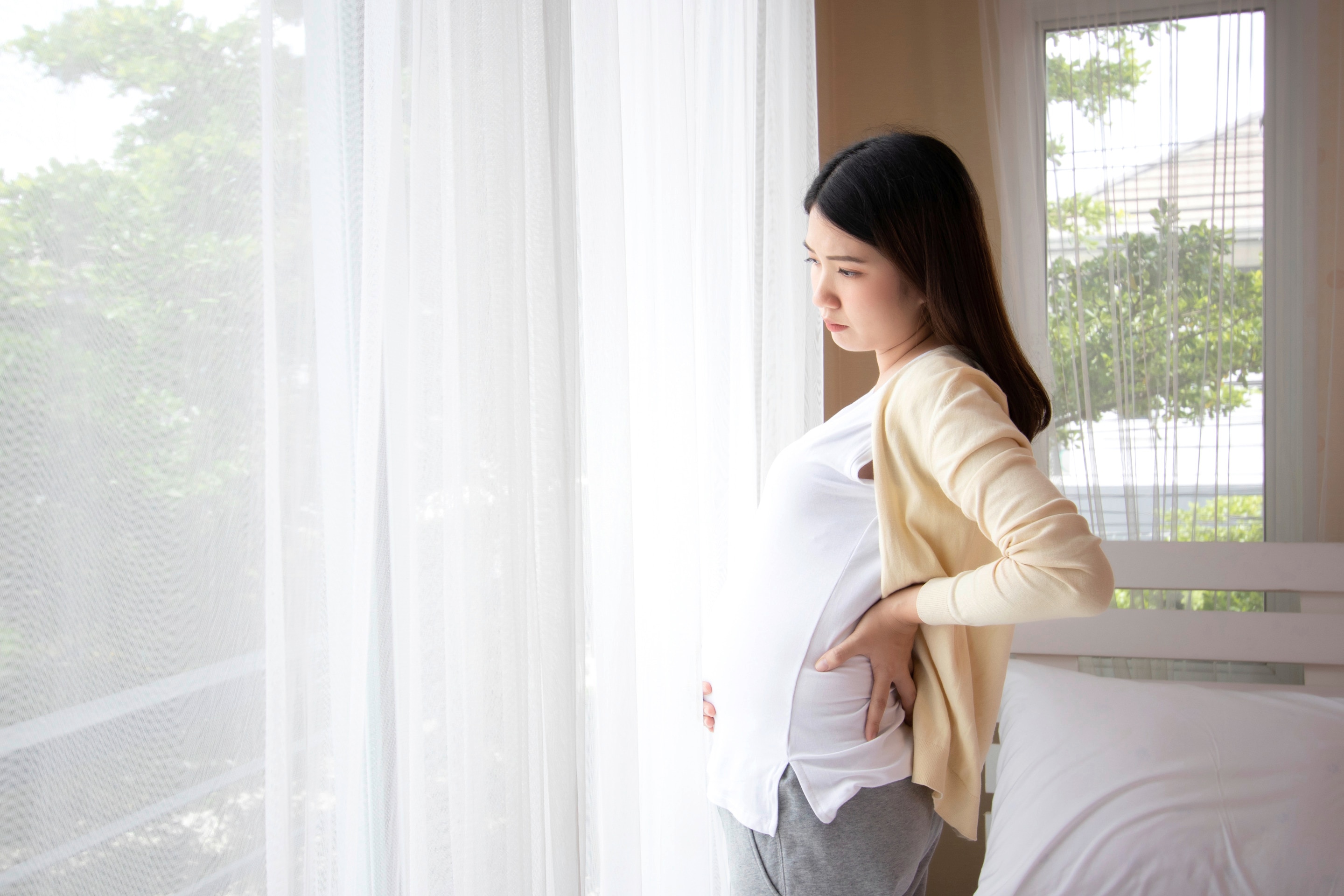 Wanita asia yang sedang hamil trimester 3 berdiri di dekat jendela, memegangi perut bawah yang sakit