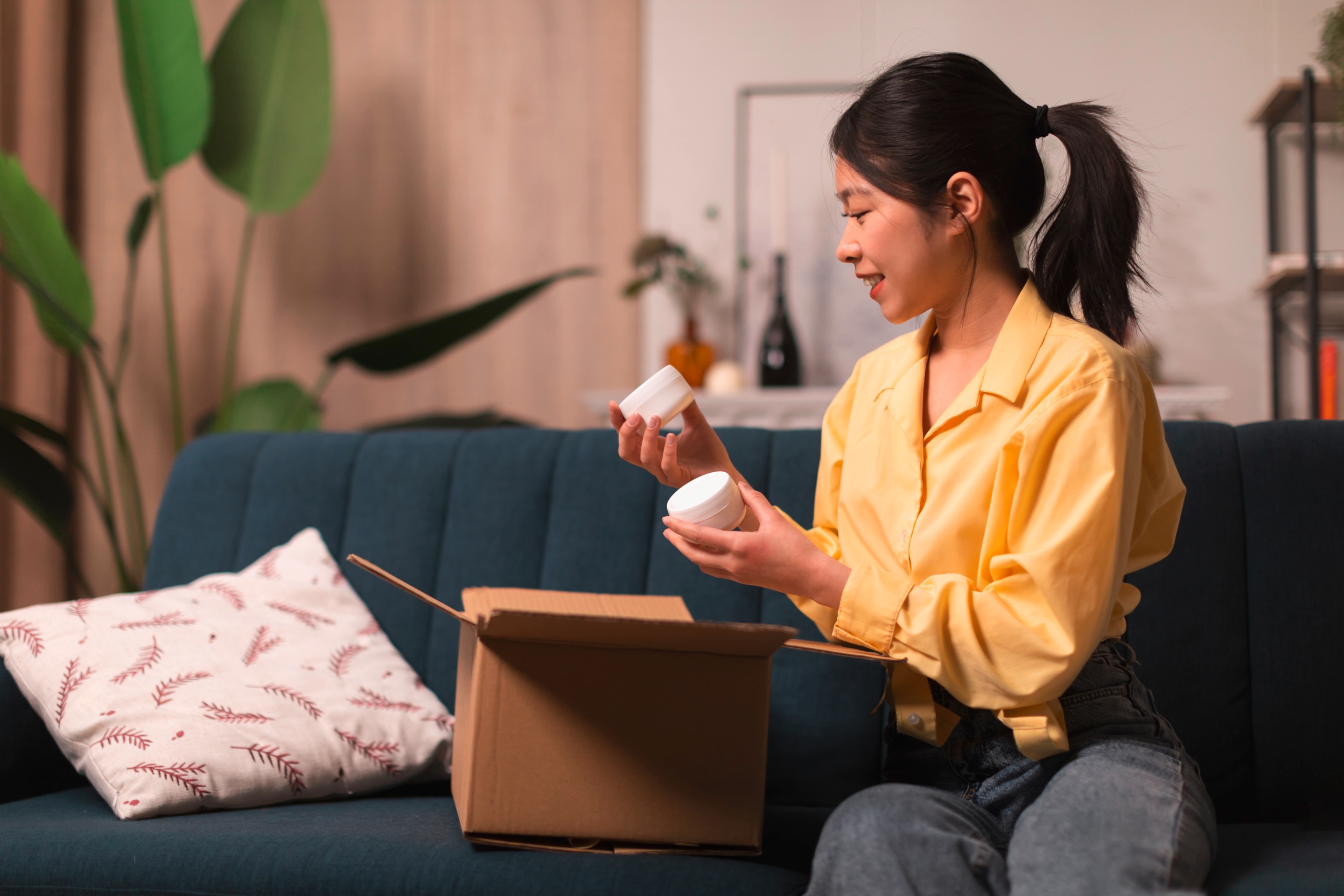 Asian woman opening box of skincare products on sofa.