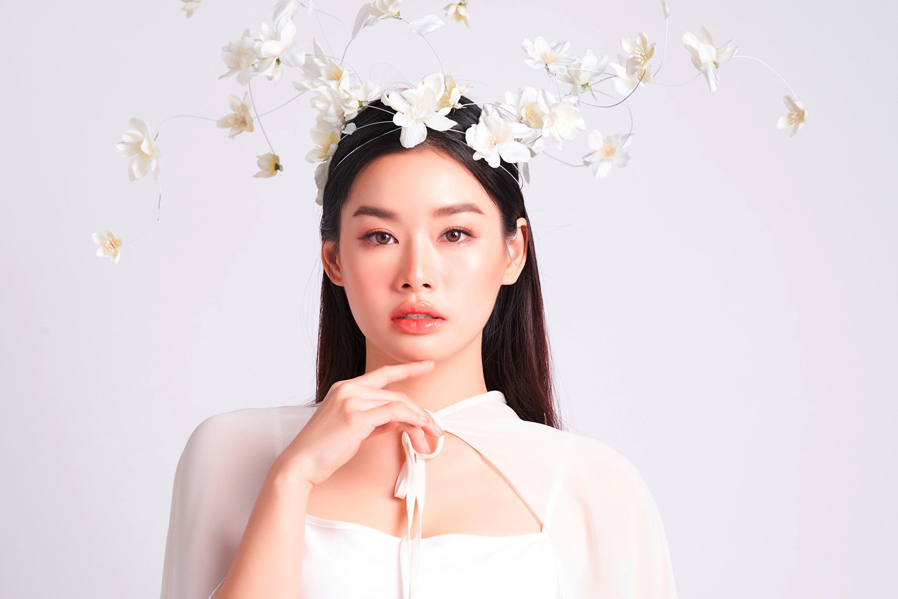 ผู้หญิงเอเชีย ผมยาวตรง บนหัวตกแต่งด้วยกิ่งดอกไม้สีขาว สวมชุดสีขาว นิ้วชี้แตะคาง
