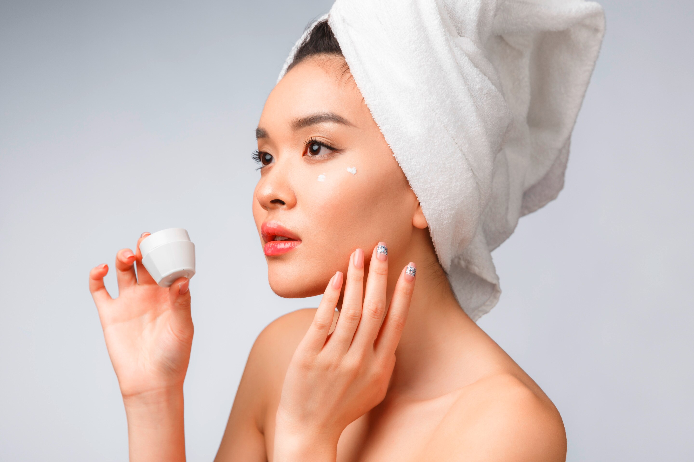 ผู้หญิงเอเชีย ผ้าขนหนูพันหัว มือถือกระปุกครีมสีขาว อีอมือแตะข้างคาง มีจุดครีมบนแก้ม