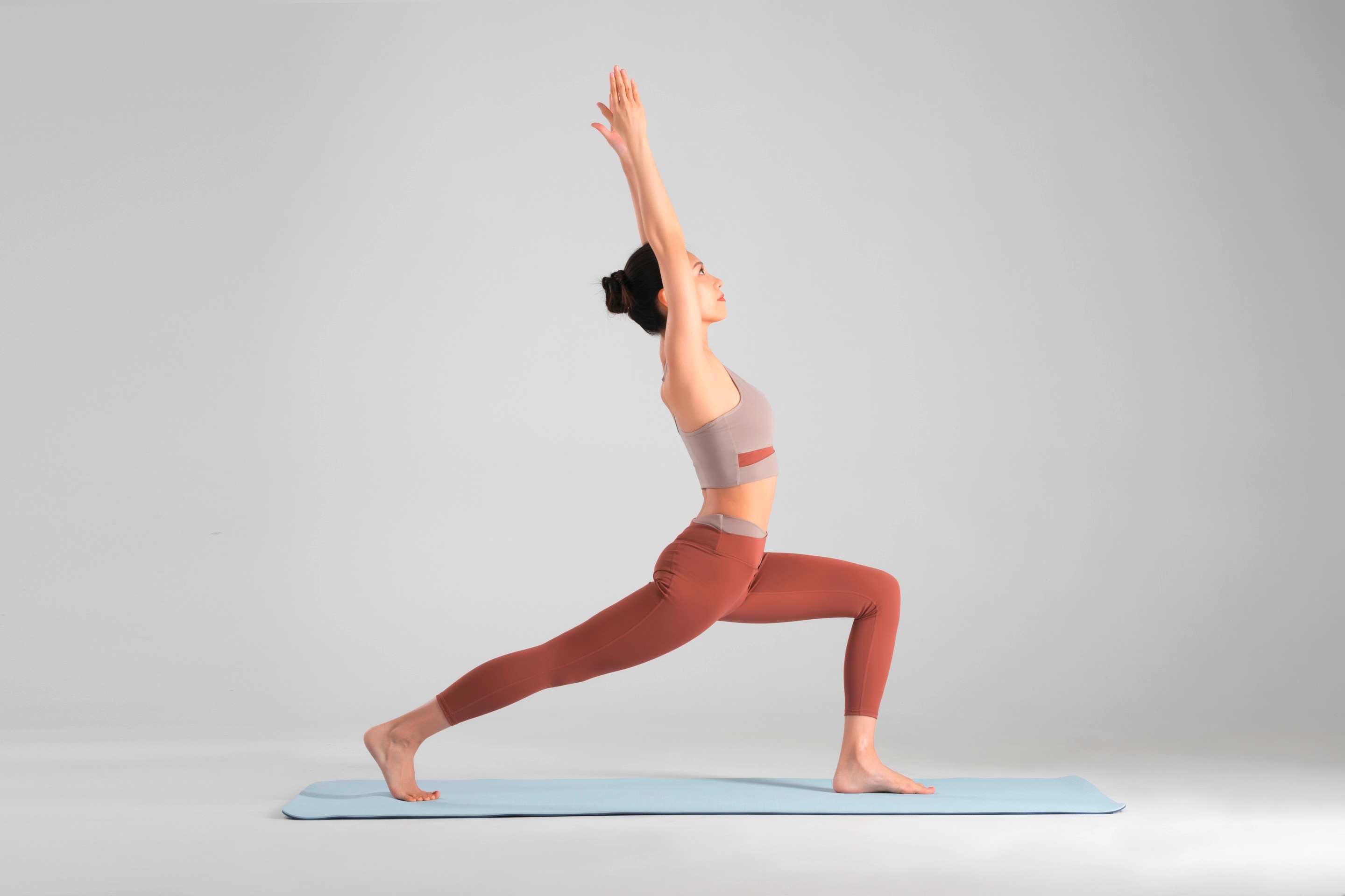 ผู้หญิงเอเชีย รวบผมมวย ทำโยคะท่านักรบบนเสื่อโยคะสีฟ้า สวมชุดออกกำลังกายสีน้ำตาล