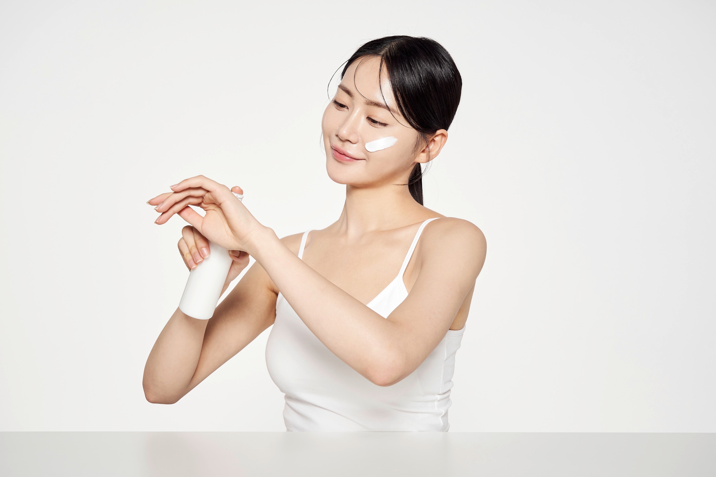 ผู้หญิงเอเชีย รวบผมมัดต่ำ กดเนื้อครีมจากขวดลงบนมือ ครีมขาวป้ายที่แก้ม สวมสายเดี่ยวสีขาว