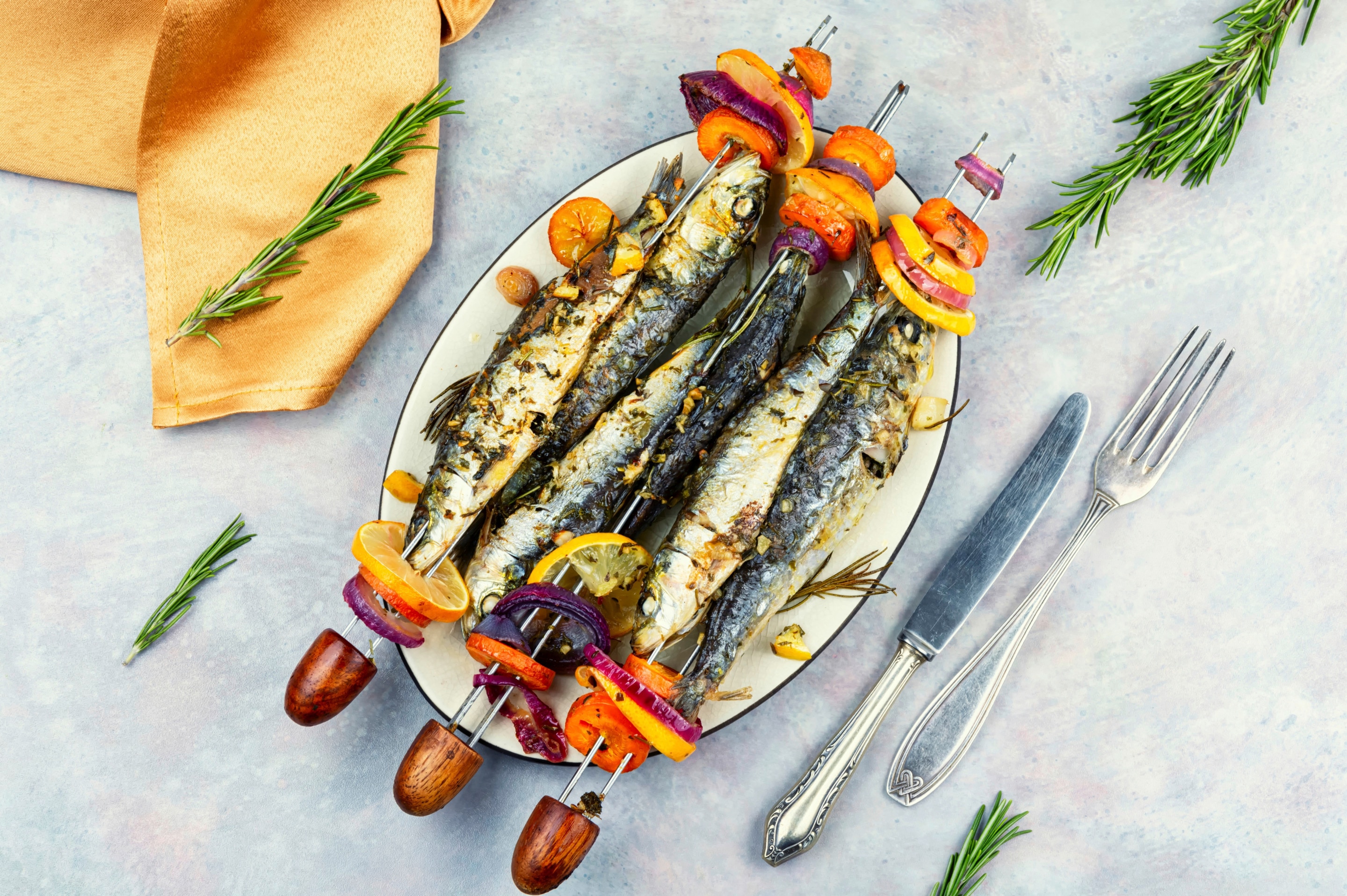 ปลาซาร์ดีนย่าง วางบนจานขาว เสียบปลาและผักสลับกัน ผ้าเช็ดปากสีส้ม ก้านโรสแมรี่ มีดกับส้อมวางด้านข้าง 