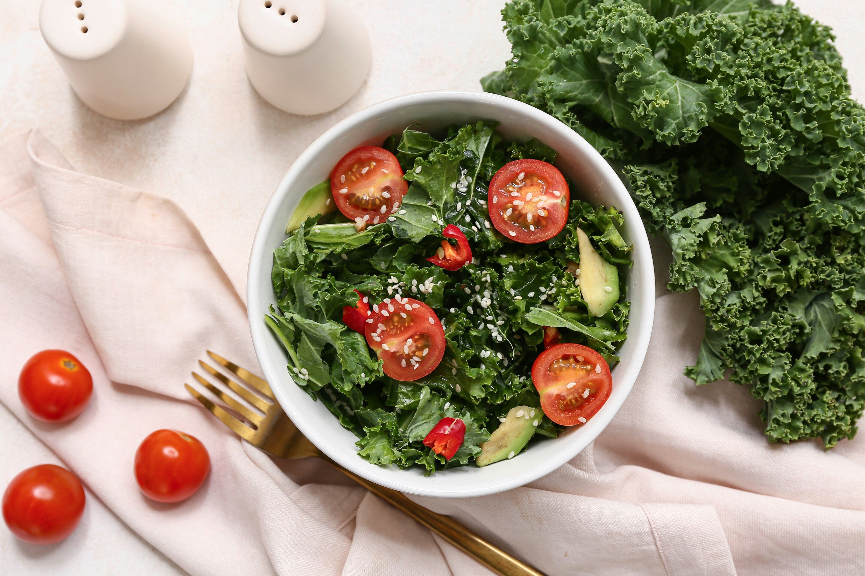 สลัดเคลในชามขาว วางบนผ้า ผักเคล มะเขือเทศเชอร์รี่ 3 ลูก ส้อมสีทอง ขวดใส่เครื่องปรุงสีขาว 2 ขวด