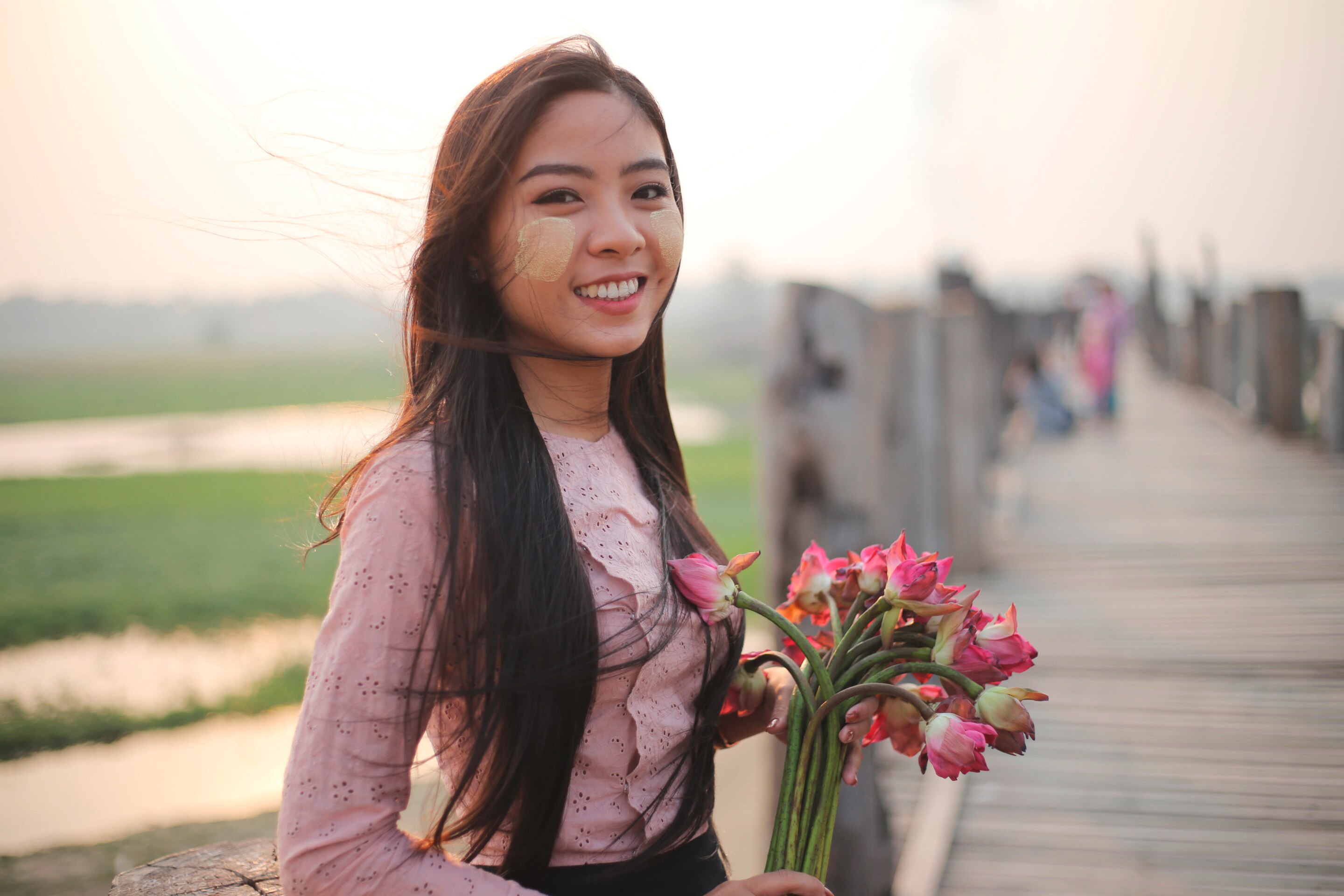 ผู้หญิงเอเชีย ผมตรงยาว ป้ายทานาคาบนแก้ม ยืนยิ้มบนสะพานไม้ ใส่เสื้อลายฉลุสีชมพู ถือช่อดอกบัวสีชมพู