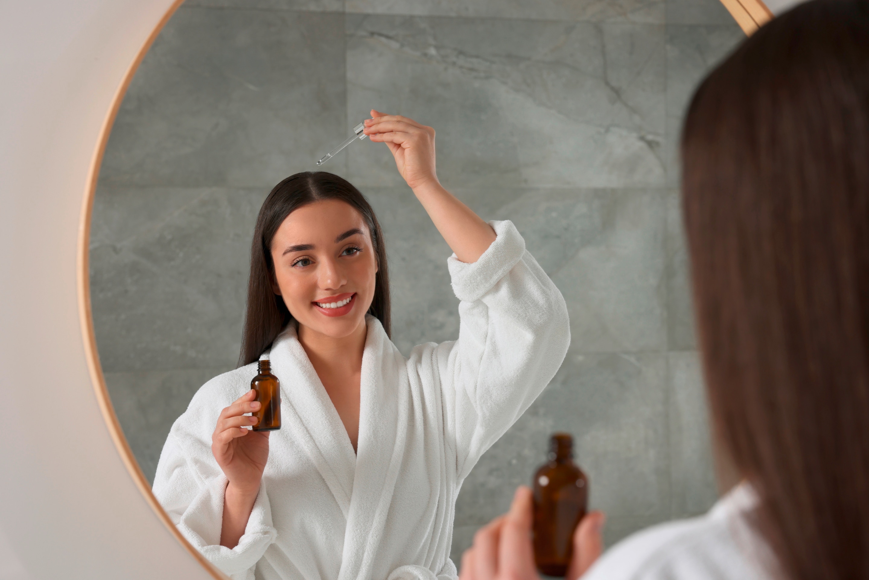 ผู้หญิงฝรั่ง ผมตรงยาวแสกกลาง สวมเสื้อคลุมอาบน้ำ ยืนส่องกระจก ถือขวดและดรอปเปอร์หยดลงบนหัว