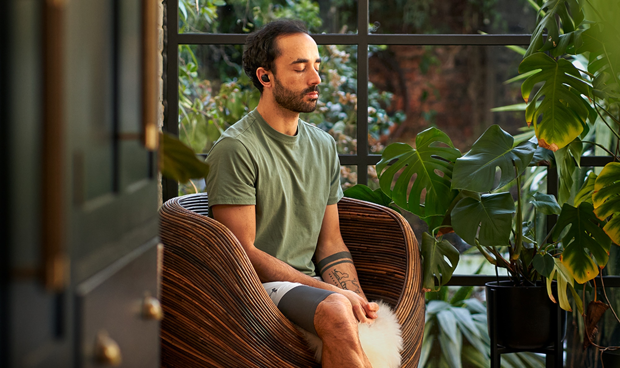 Man with eyes closed, meditating in a wicker chair, surrounded by houseplants and natural light