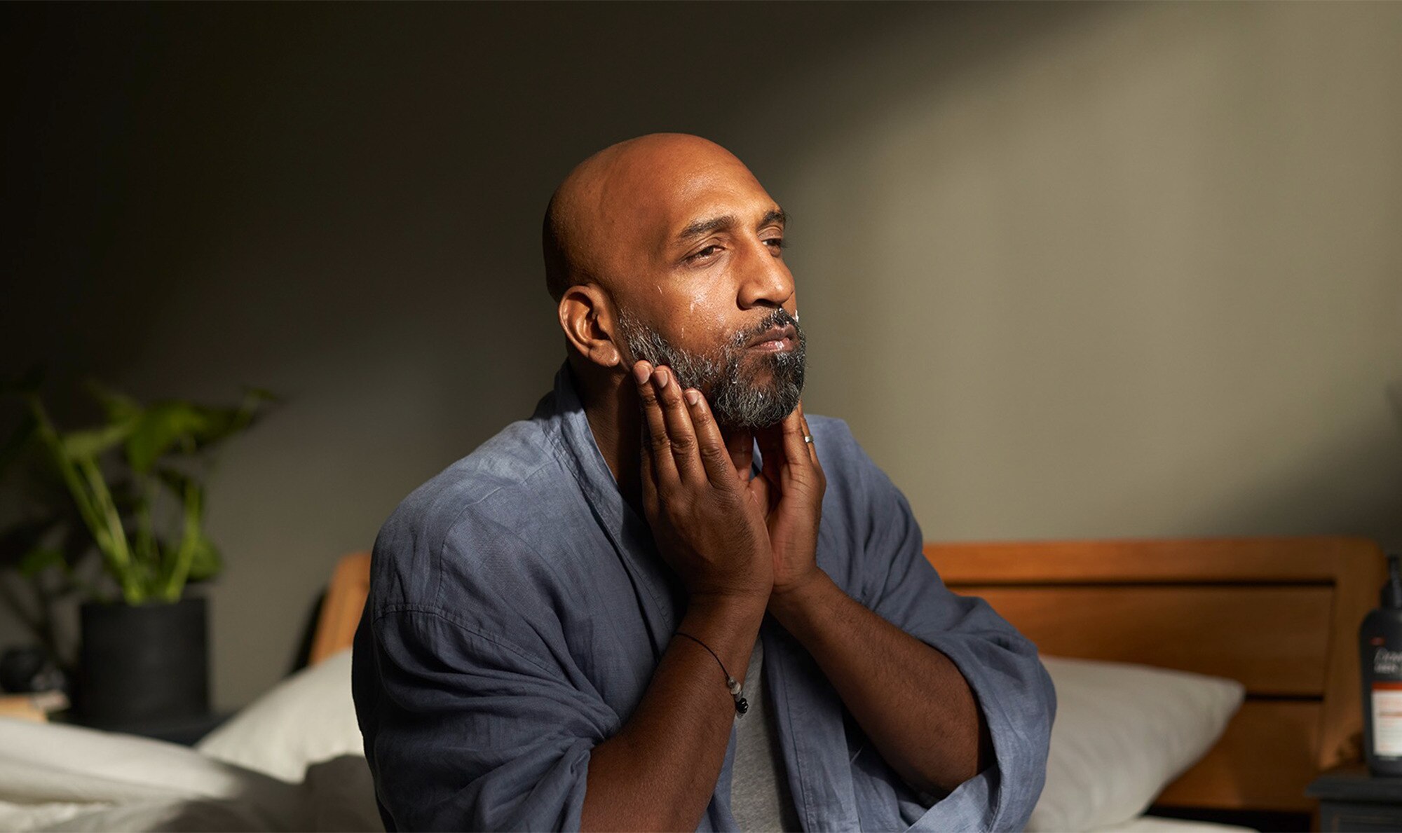 Man touching his face with both hands, sitting on the edge of a bed