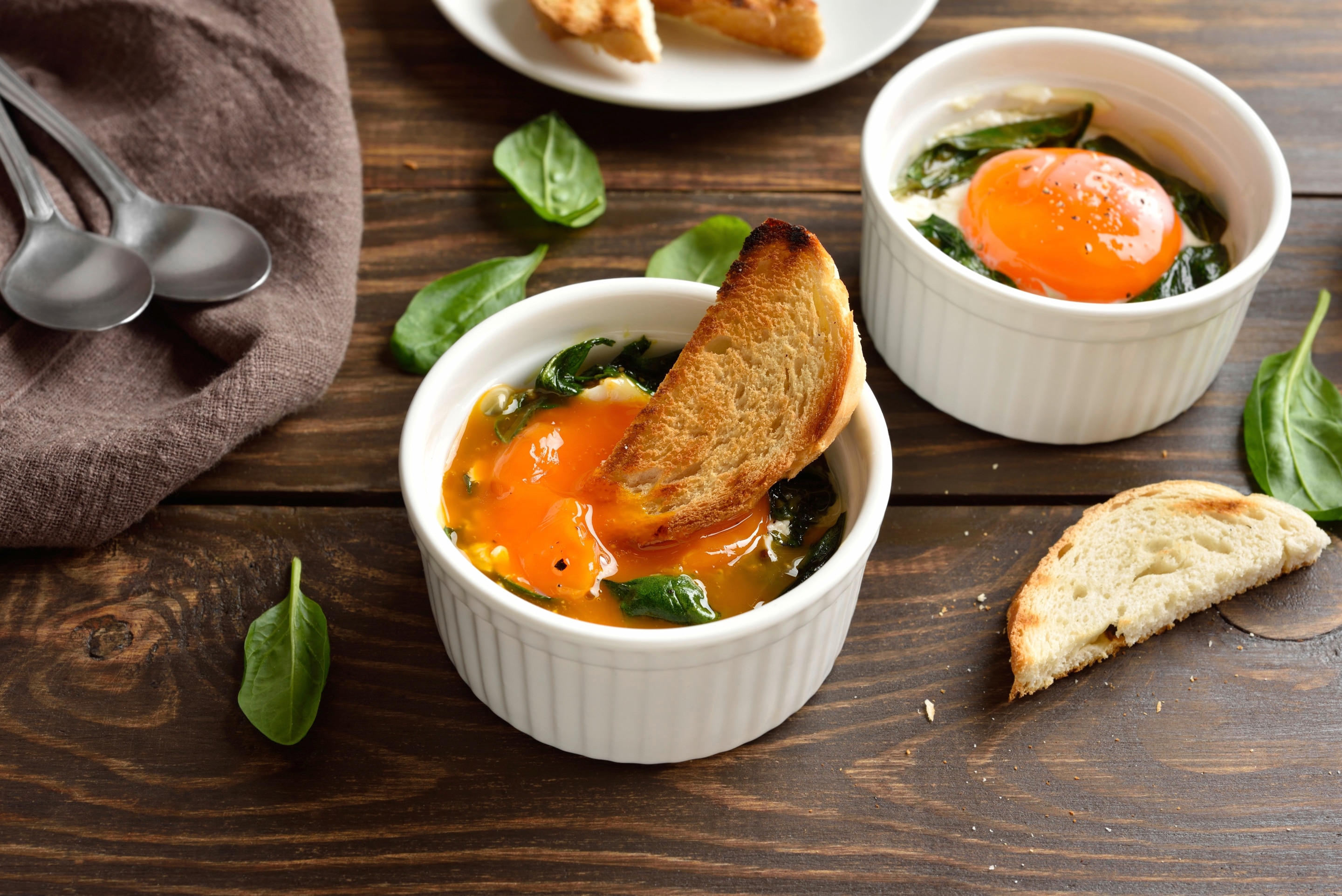 ไข่แดงอบผักโขม ขนมปังปิ้งหั่น ผักโขมสดโรยบนโต๊ะไม้ ช้อนสองคันบนผ้าสีน้ำตาล