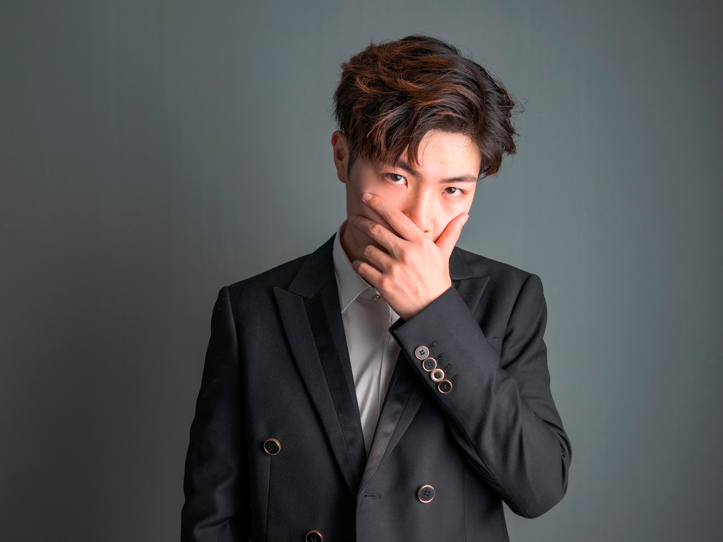 ผู้ชายเอเชีย ตัดผมทรงทูบล็อก ผมทำสีไฮไลต์ มือกุมปากและคาง สวมเชิ้ตขาวทับดวยเบลเซอร์สีดำ