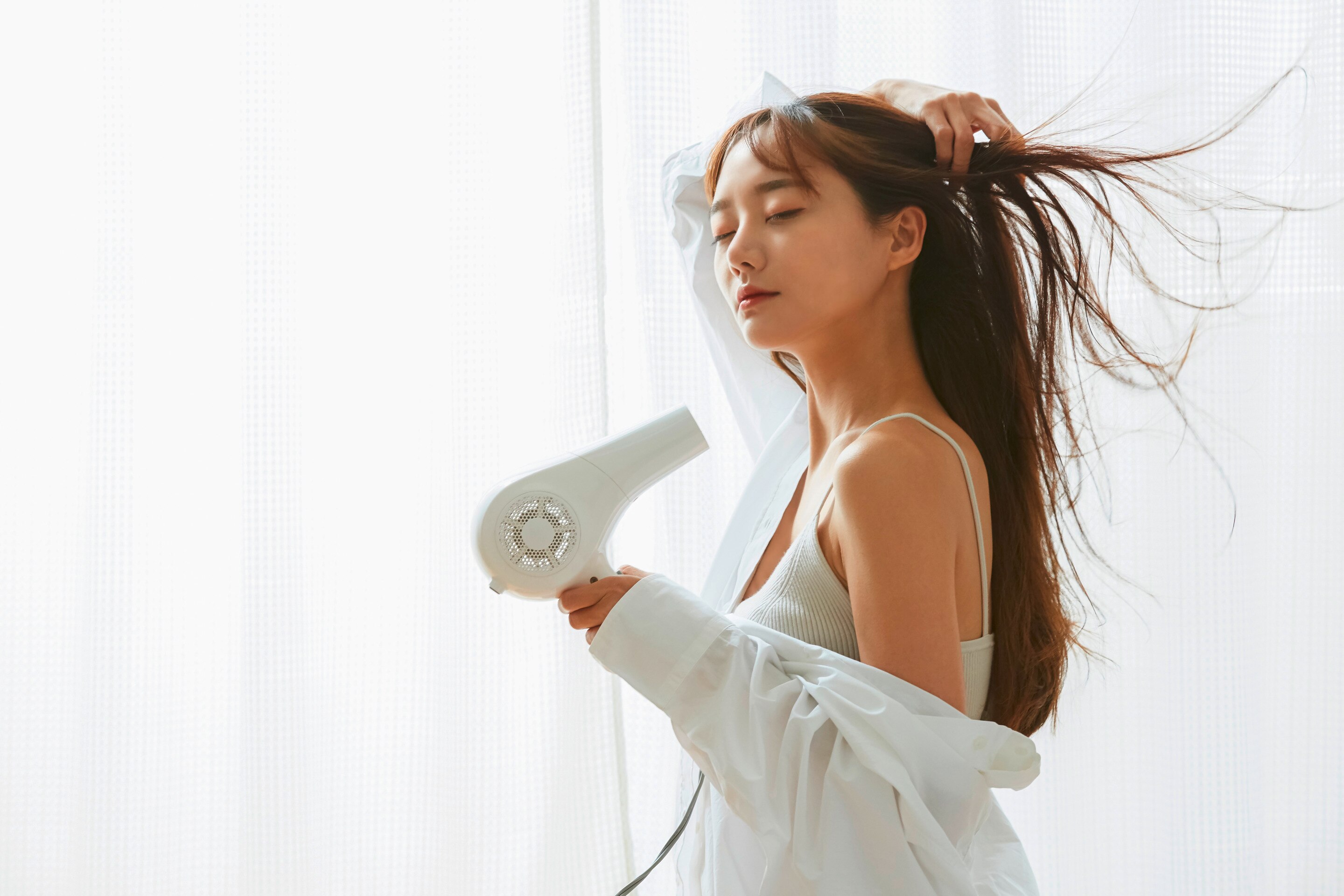 ผู้หญิงเอเชีย ผมตรงยาวพลิ้วตามแรงลมจากไดร์ สวมเชิ้ตสีขาวเปิดไหล่