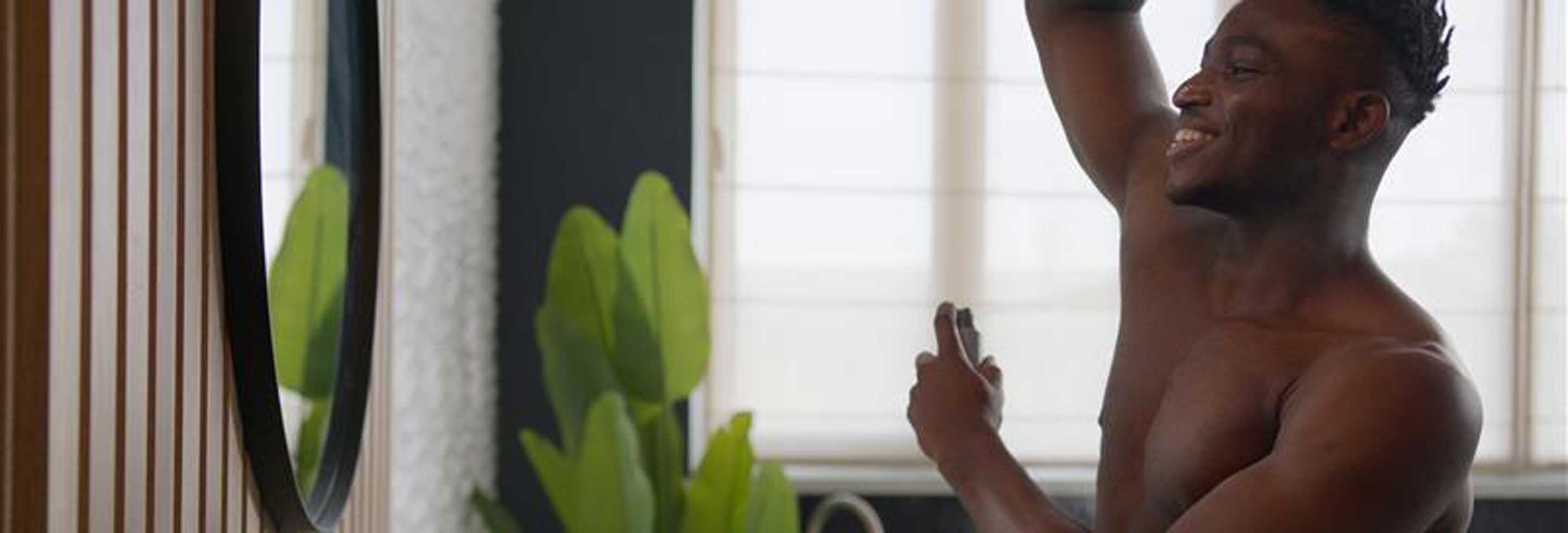 Smiling shirtless man applying spray deodorant in front of a bathroom mirror
