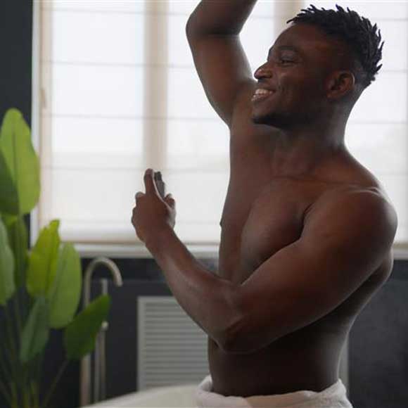 Thumbnail image of smiling shirtless man applying spray deodorant in front of a bathroom mirror 