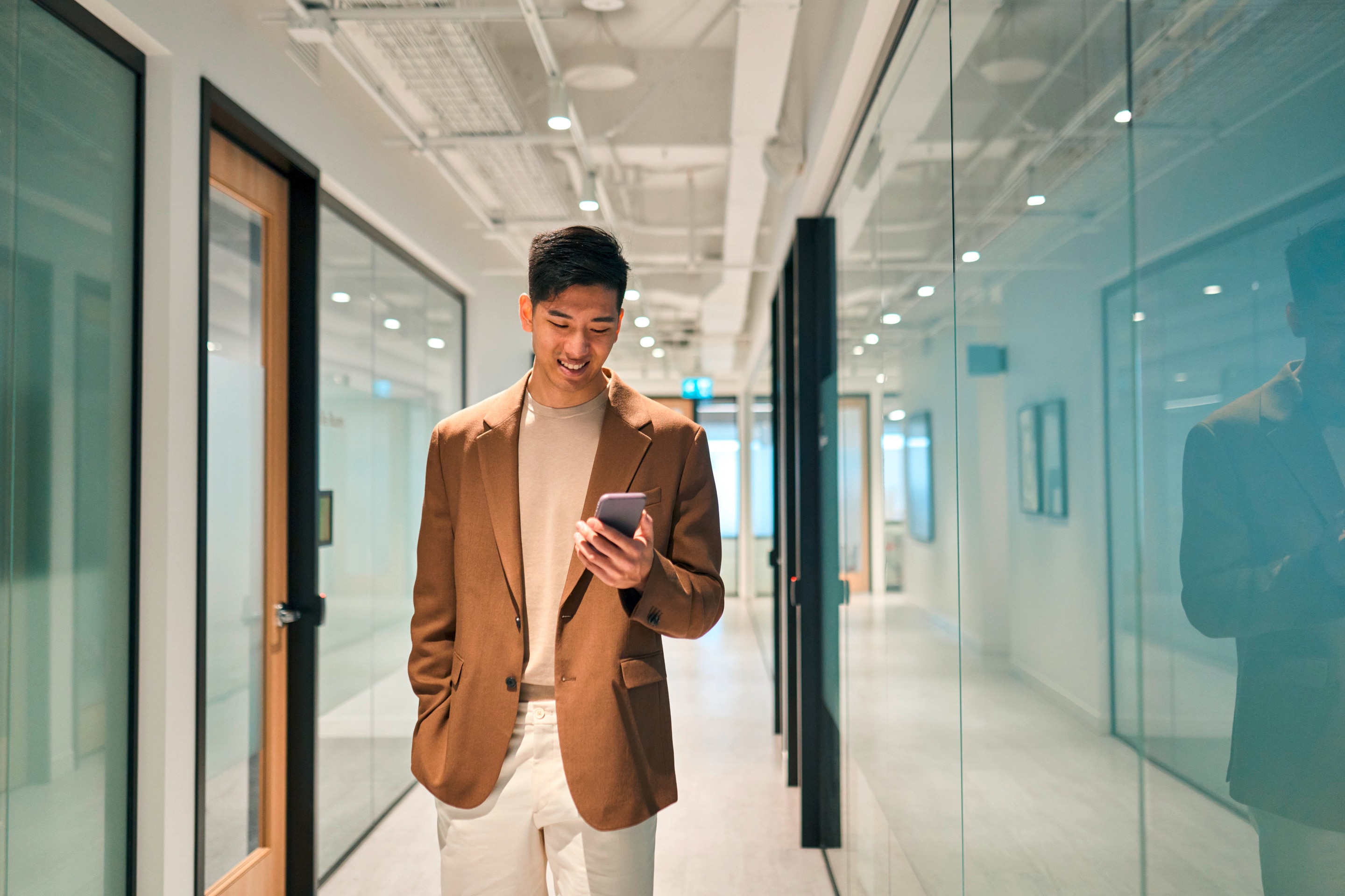Asian man in brown blazer smiling at his phone.