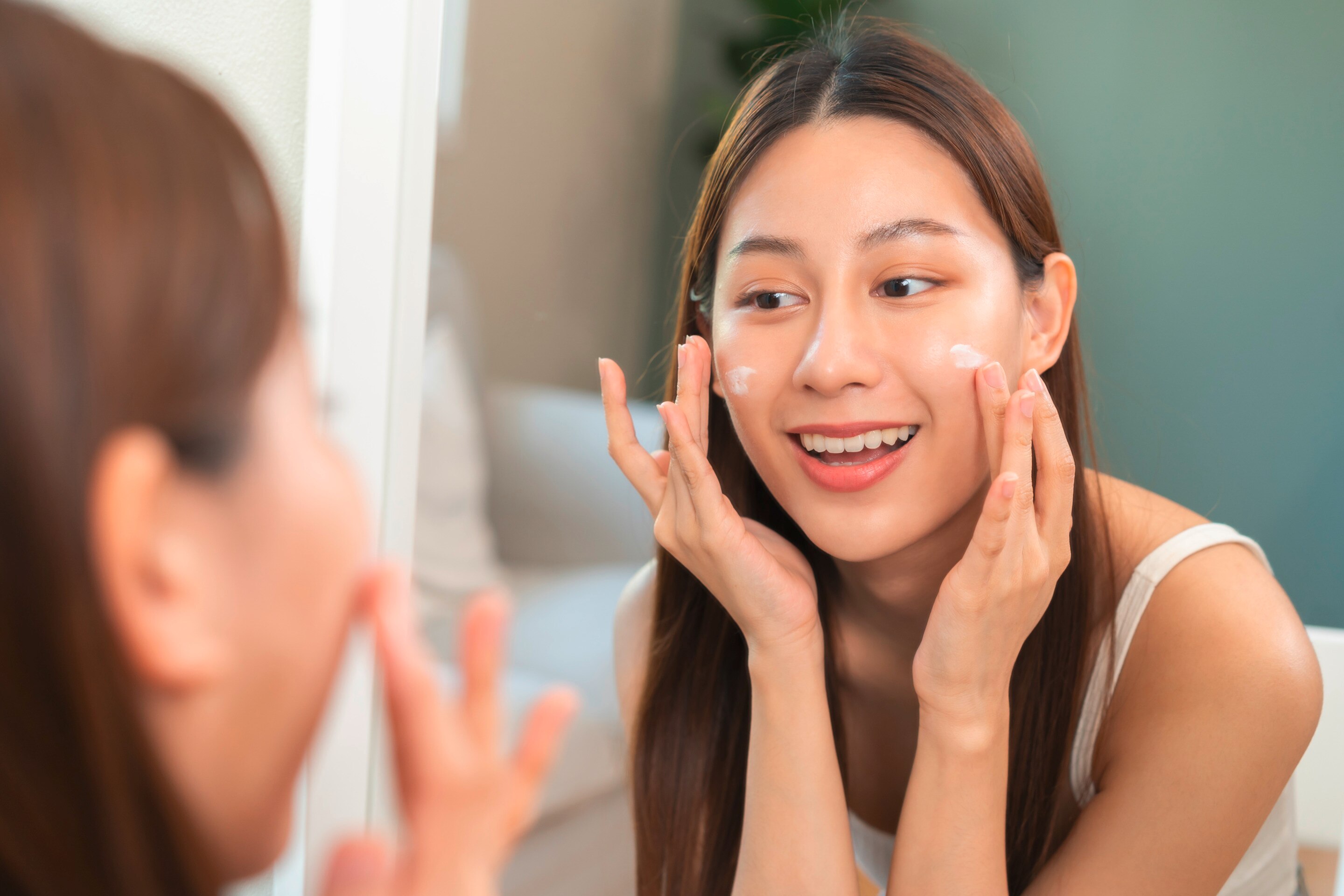 A woman happily applies moisturizer in front of a mirror.