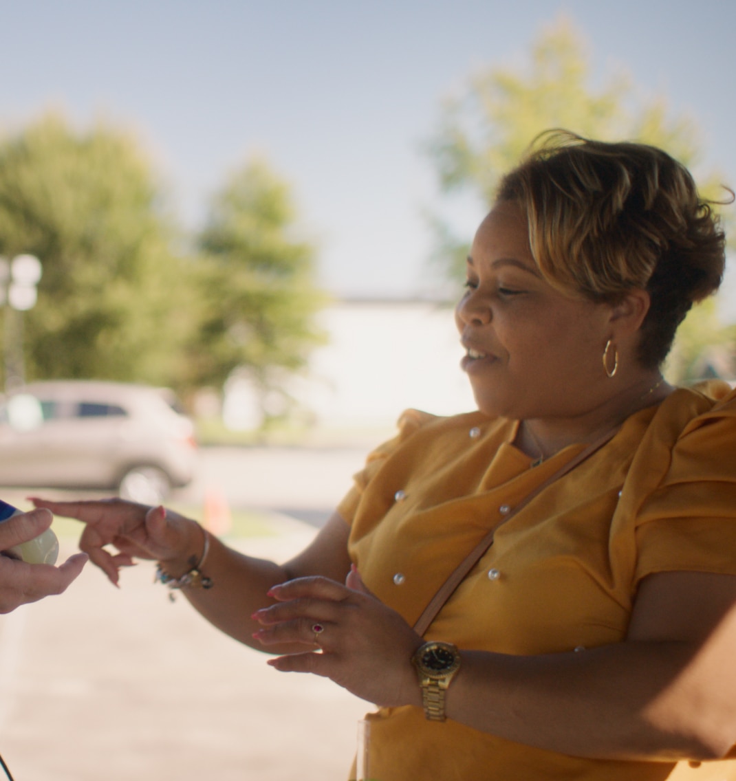 A person wearing a bright yellow top with pearl-like accents is interacting with another individual outdoors near parked cars