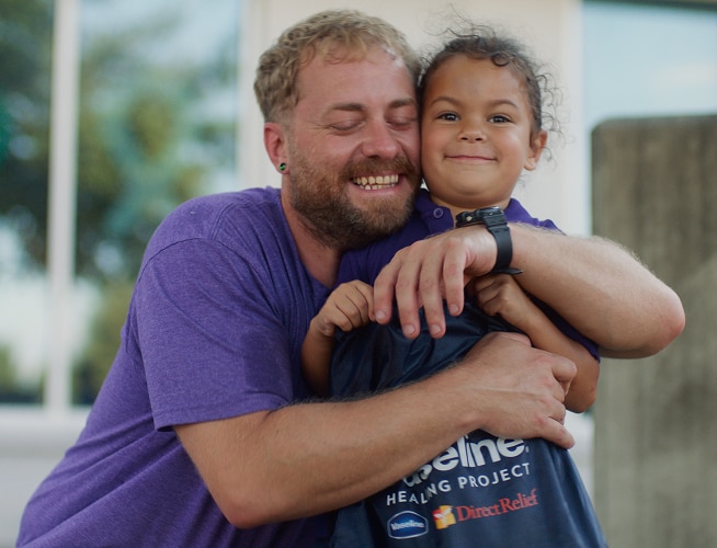 Adult hugging a child holding a Vaseline Healing Project souvenir as they smile happily