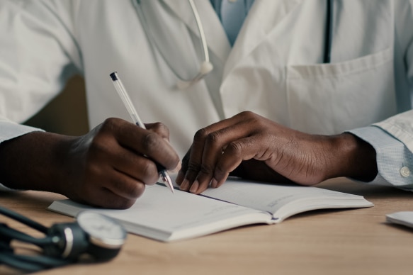 A doctor writing notes in a medical journal with a stethoscope placed on the desk