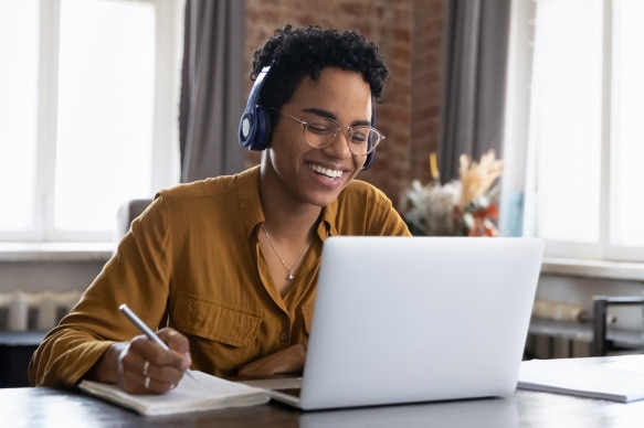 A person taking notes while attending an online class on a laptop with headphones, representing a modern remote learning setup