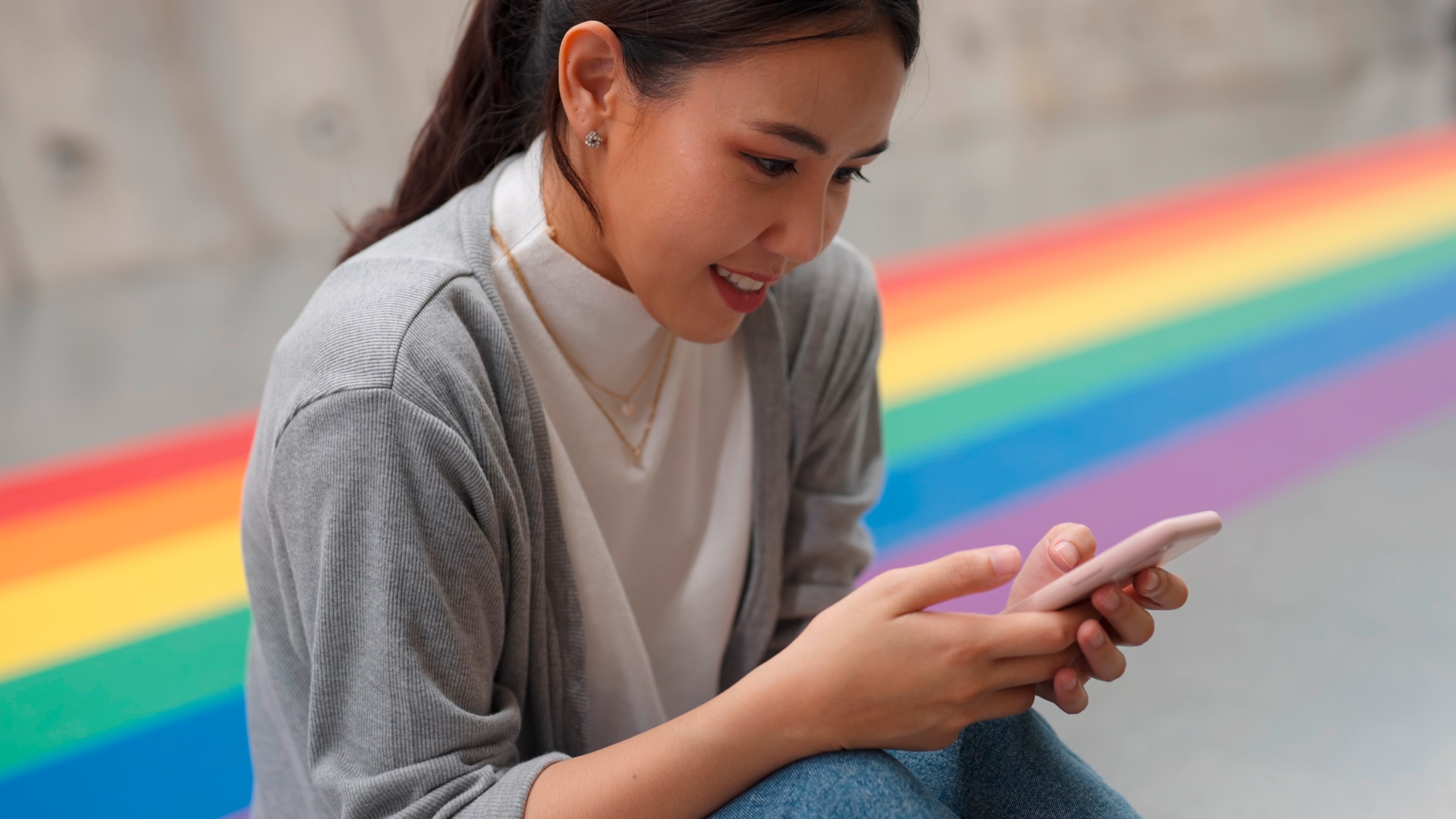 Queer Asian woman scrolling on her phone with Pride flag colors behind her.