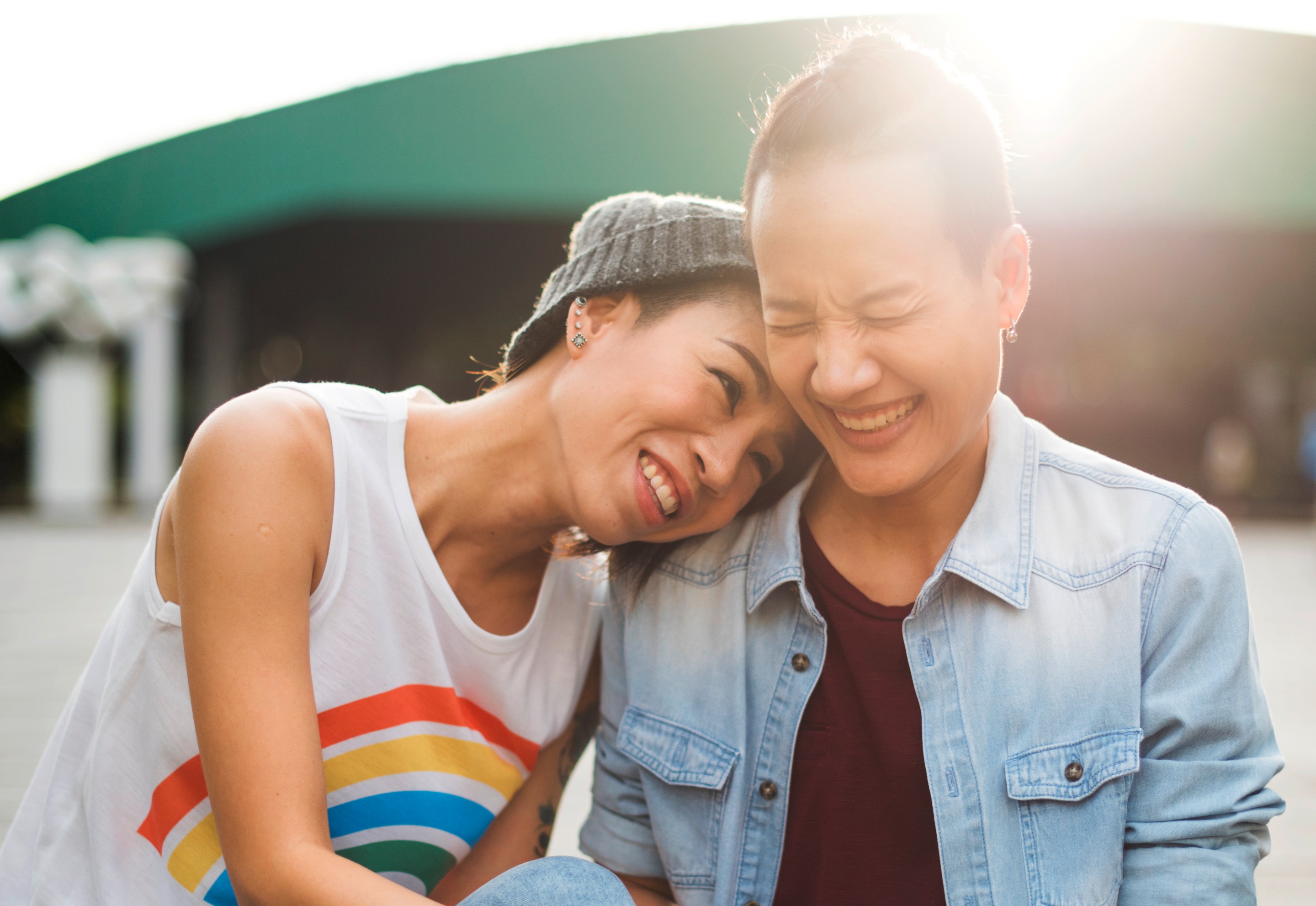 Two young queer Asian women laughing and sitting outdoors.